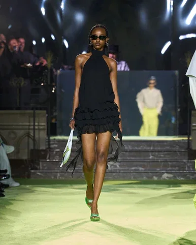 Une modèle marche sur un podium lors d'un défilé de mode, portant une robe noire courte, des lunettes de soleil et des chaussures vertes, avec un public et un animateur en arrière-plan.