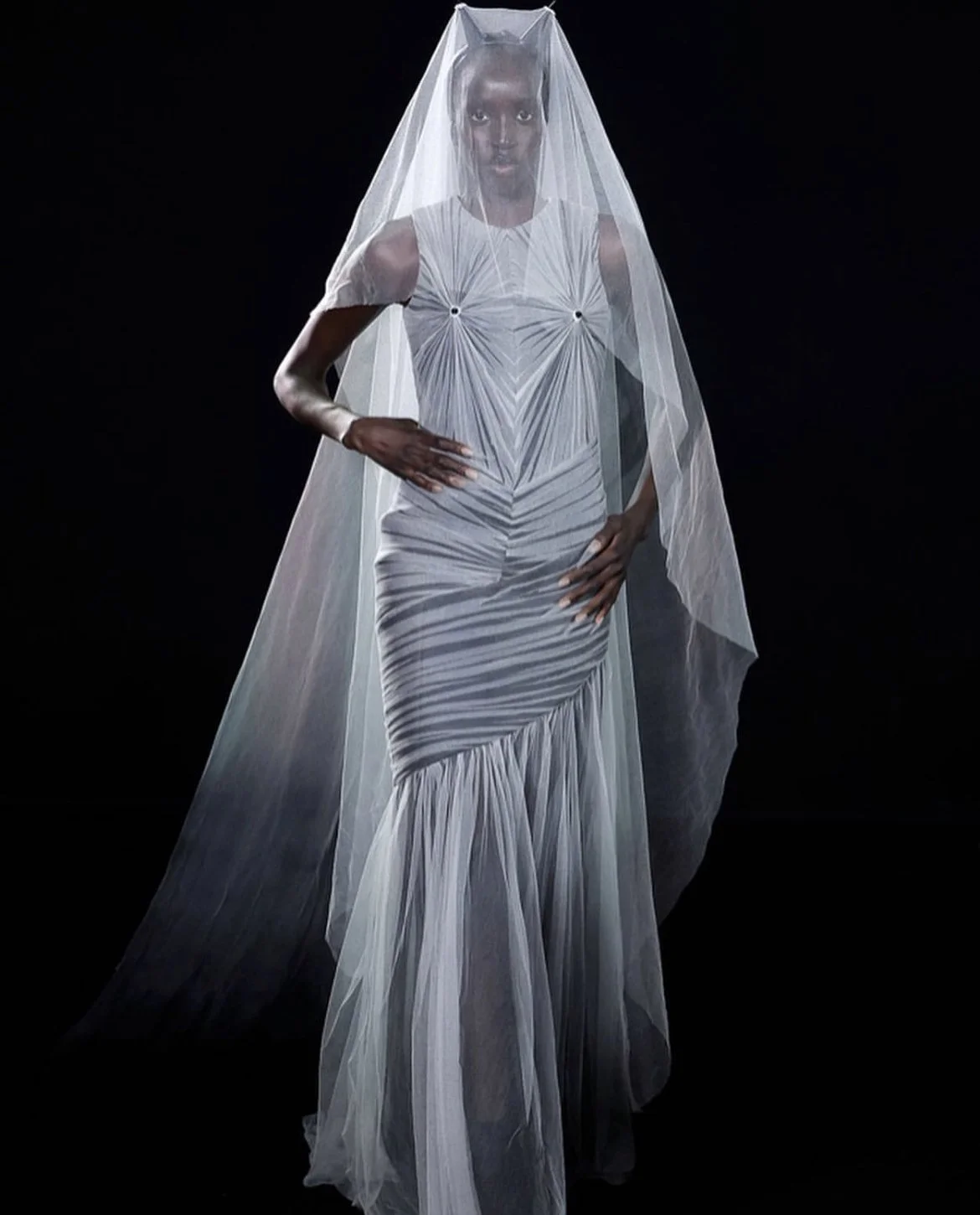 Une femme en robe blanche avec un voile transparent sur la tête, posant contre un fond noir.