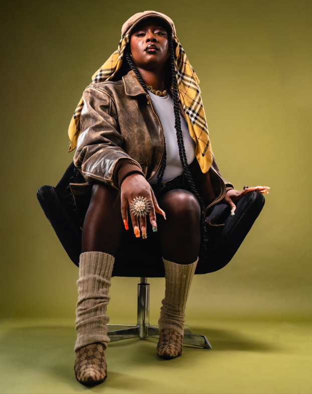 Une femme assise sur une chaise noire, portée par une version plus large, portant un chapeau, une veste en cuir, une écharpe à carreaux, des chaussettes épaisses et des bottines à motif de serpent, avec des bijoux voyants.