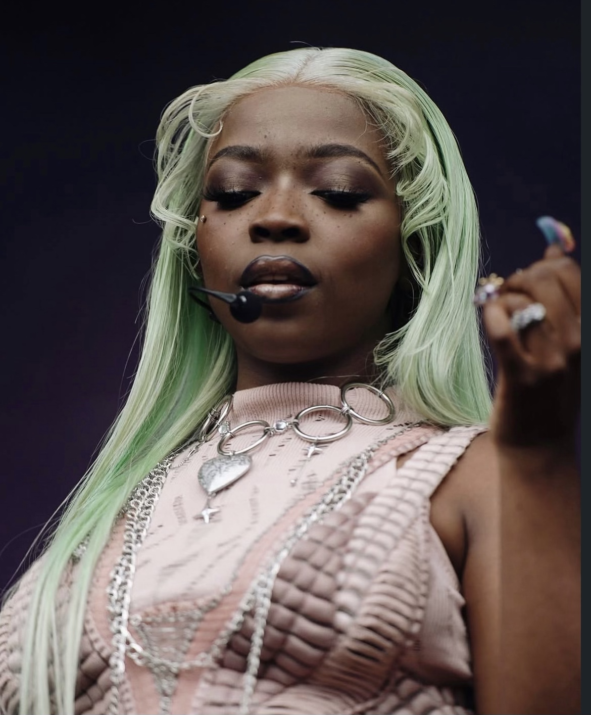 Femme avec cheveux longs verts, maquillage accentué, portant une chaîne de collier, vêtue d'une tenue rose, et un microphone près de la bouche, semblant en performance ou enregistrement.