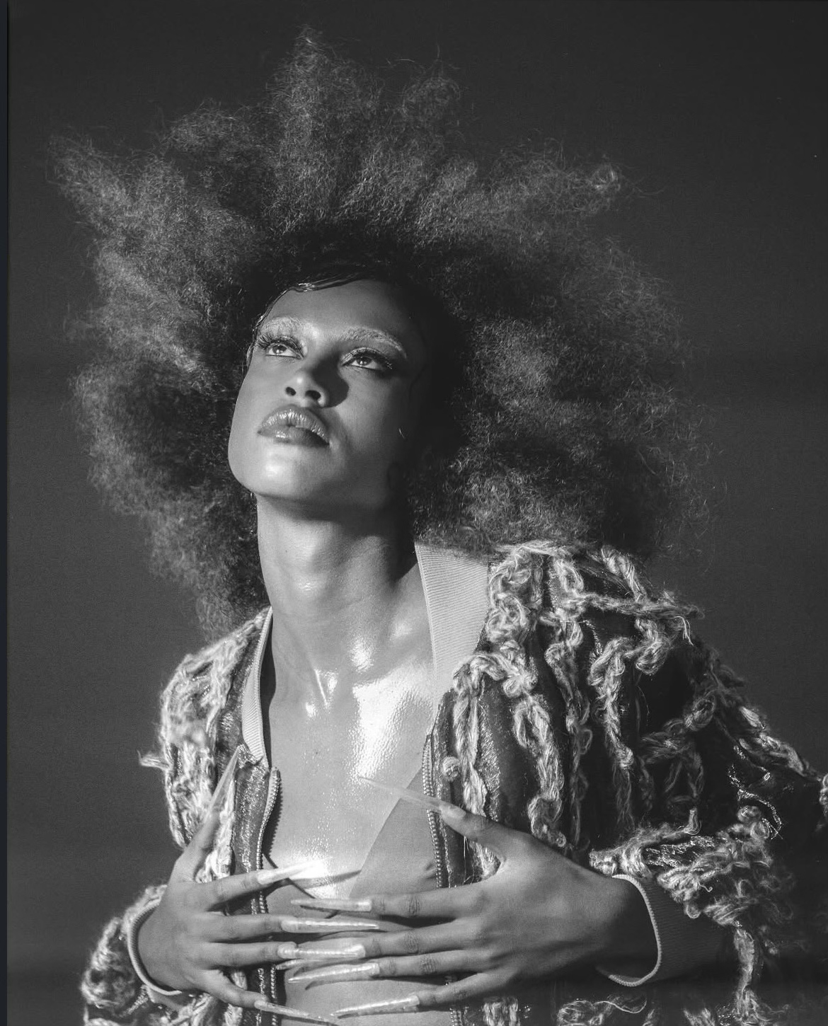 Une femme avec une coiffure volumineuse et bouclée, portant une veste en tissu avec des éléments en boucle, tenant ses mains sur sa poitrine dans une pose expressive.