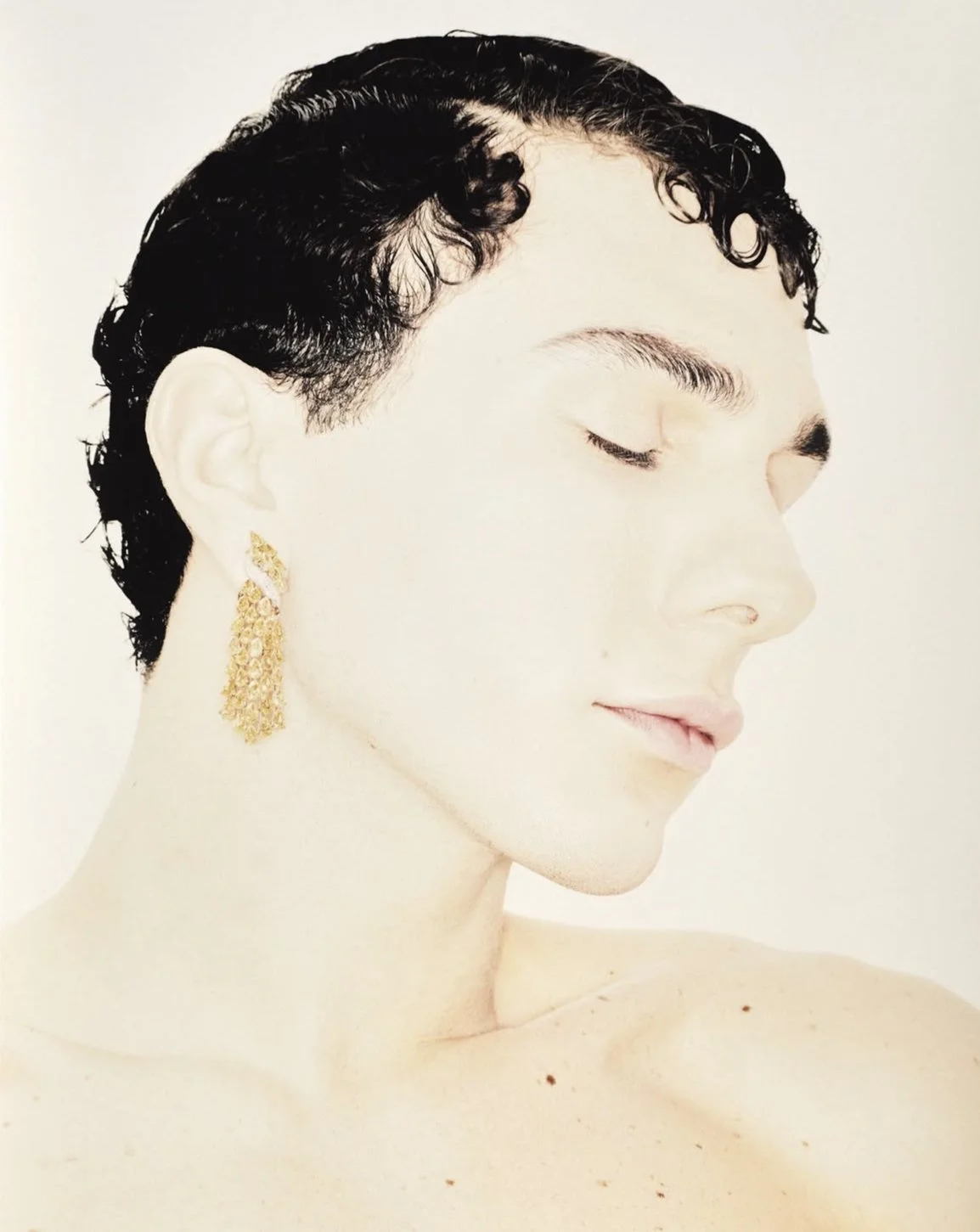 Portrait d'une femme aux cheveux courts et noirs, portant une grande boucle d'oreille dorée, vue de profil avec les yeux fermés.