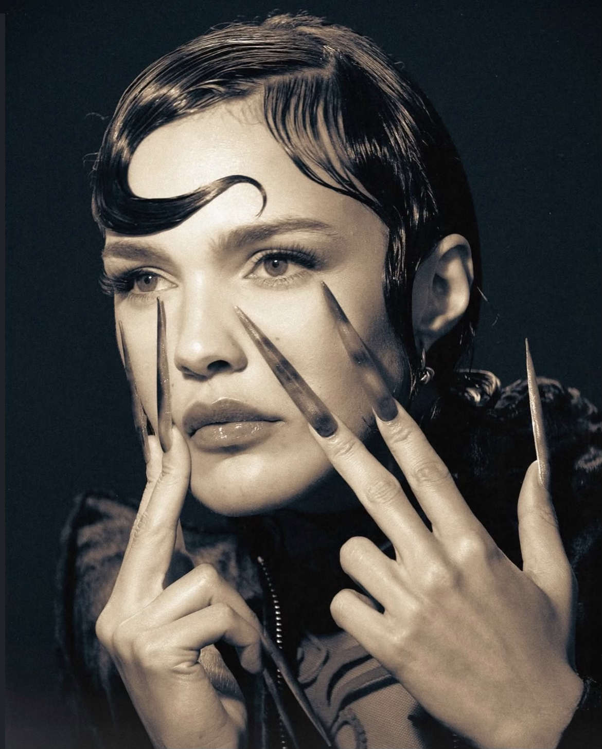 Image en noir et blanc d'une femme avec des cheveux courts, portant un maquillage léger, tenant ses mains avec des ongles longs et pointus près de son visage, avec un fond sombre.