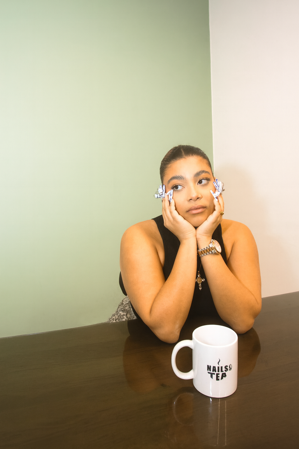 Jeune femme assise à une table, portant un haut noir, avec des rouleaux dans les cheveux, regardant pensivement à gauche, une tasse blanche avec l'inscription 'NAILS & TEA' sur la table devant elle.