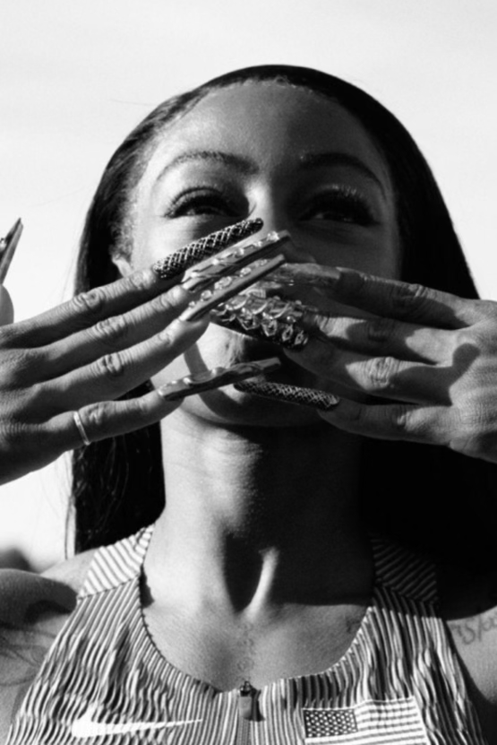 Une femme souriante avec des ongles décorés, portant une veste avec un drapeau américain, couvre la bouche avec ses mains.