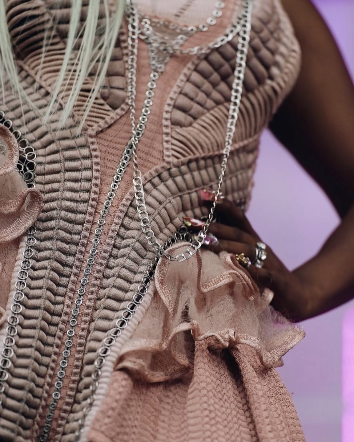 Une femme porte une robe en tissu beige avec des détails de dentelle et des chaînes en argent autour du cou, avec des bijoux et des fleurs en tissu embellissant la robe.