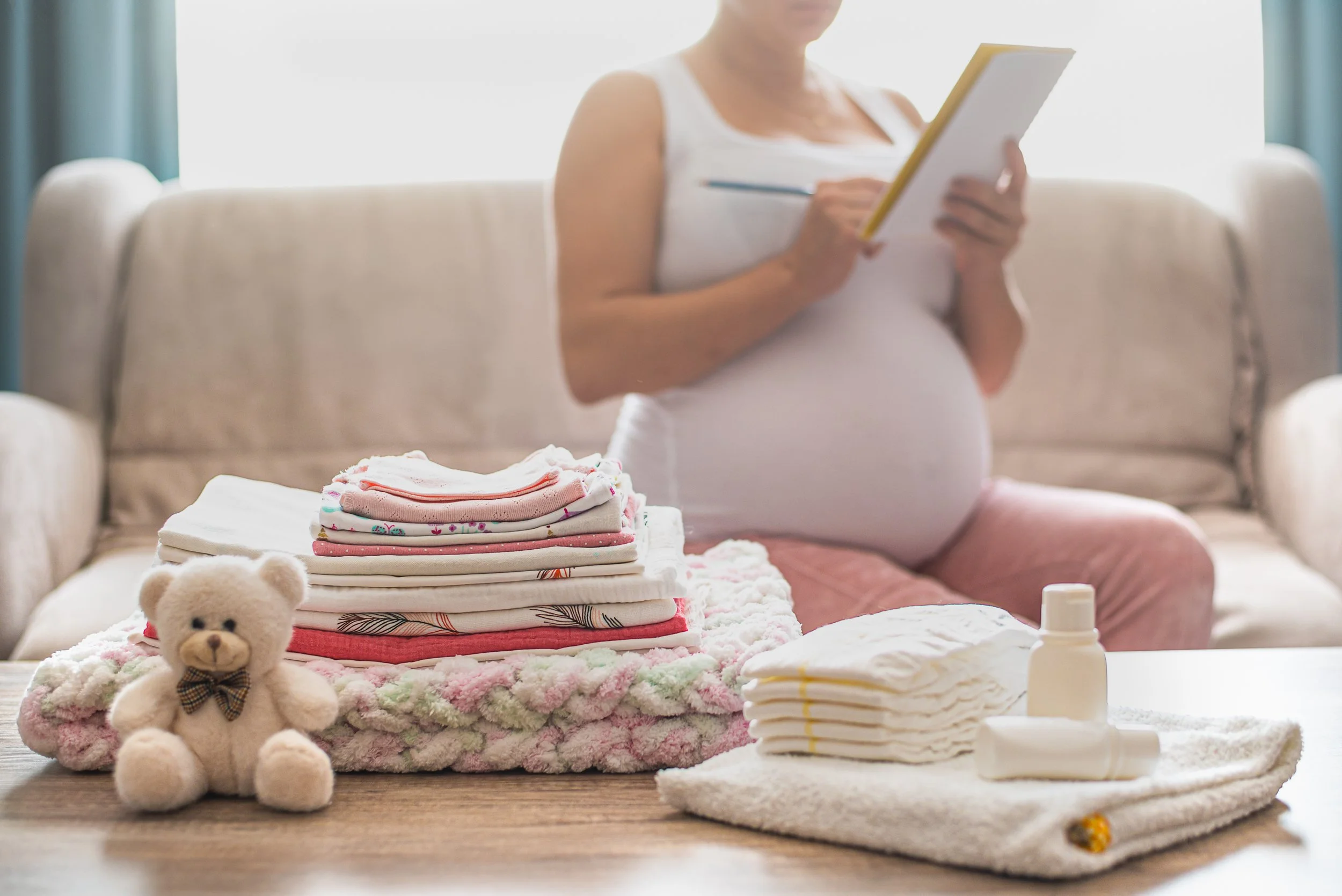 A pregnant woman prepares for her baby next to a stack of folded baby clothes placed on a fluffy blanket with a teddy bear in front, and baby lotion, shampoo, or powder bottles nearby.