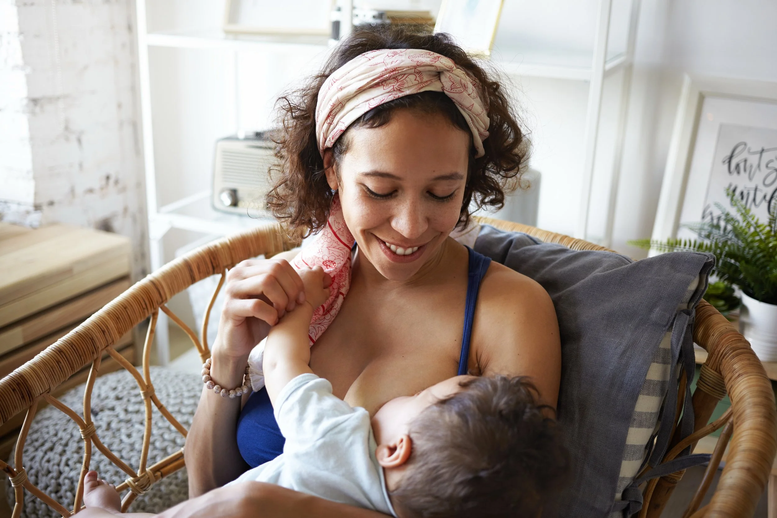 A mother with curly hair and a light-colored bandana on her head is smiling as she looks down at an older infant. She is sitting in a wicker chair indoors, with a window and framed wall art in the background.