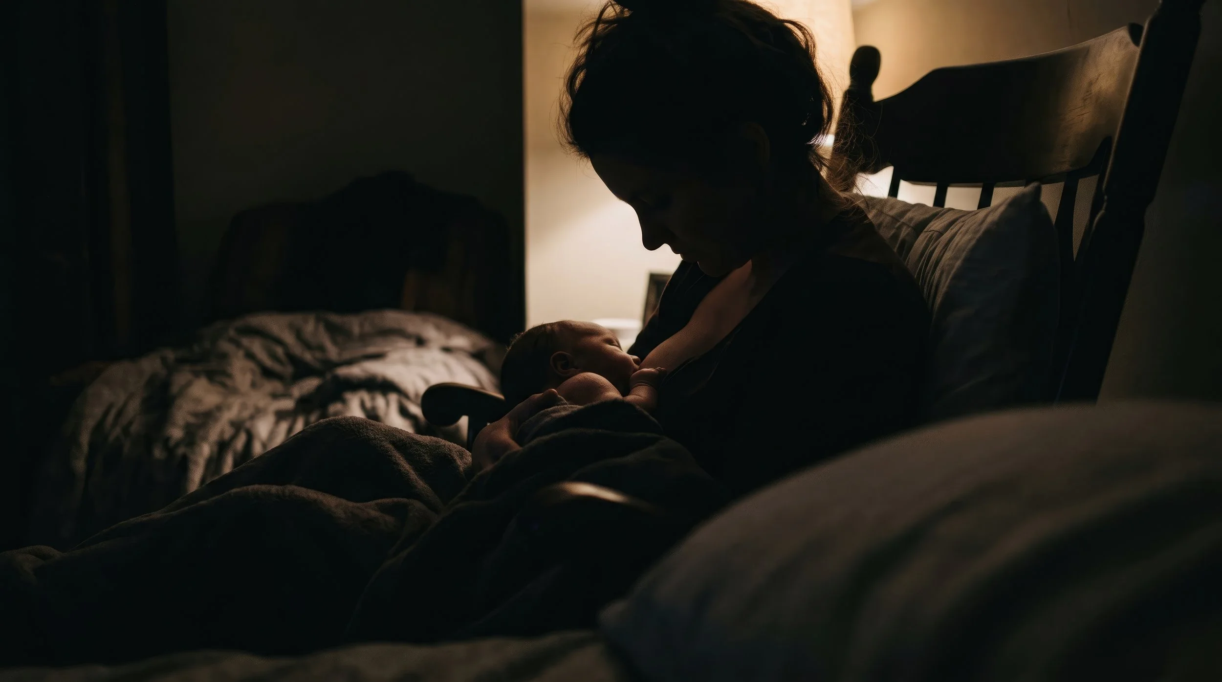 A woman sitting on a bed in a dark room, breastfeeding a newborn baby under warm lighting.