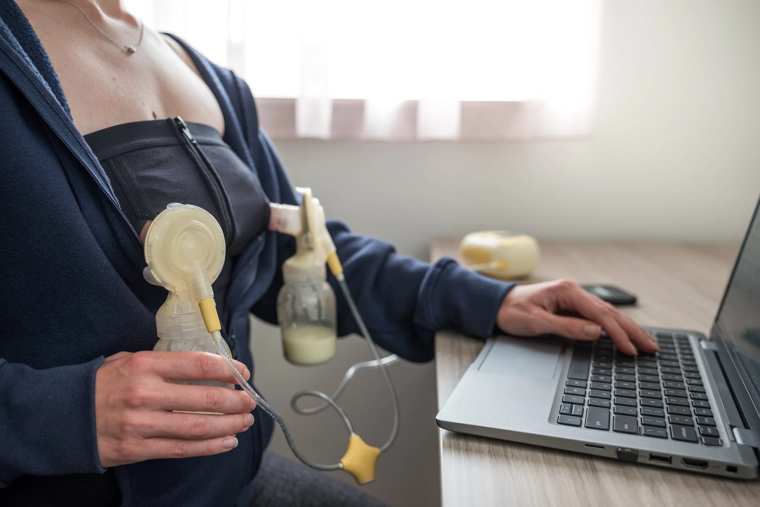 A working mother from home using a portable breast pump connected to their breast, using a laptop on a desk.