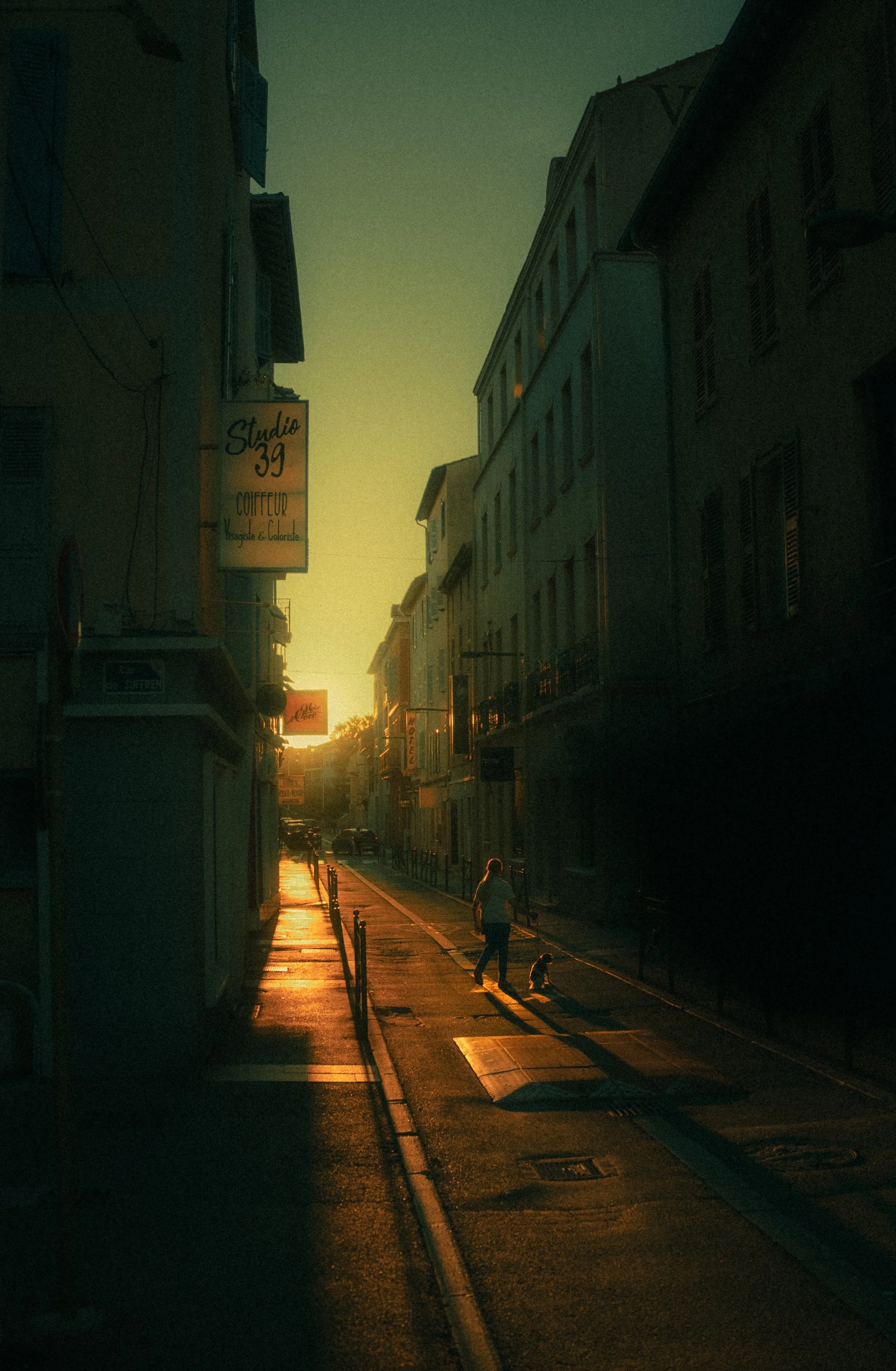 Une rue pavée au coucher du soleil avec une personne marchant un chien, entourée de bâtiments résidentiels et de commerces