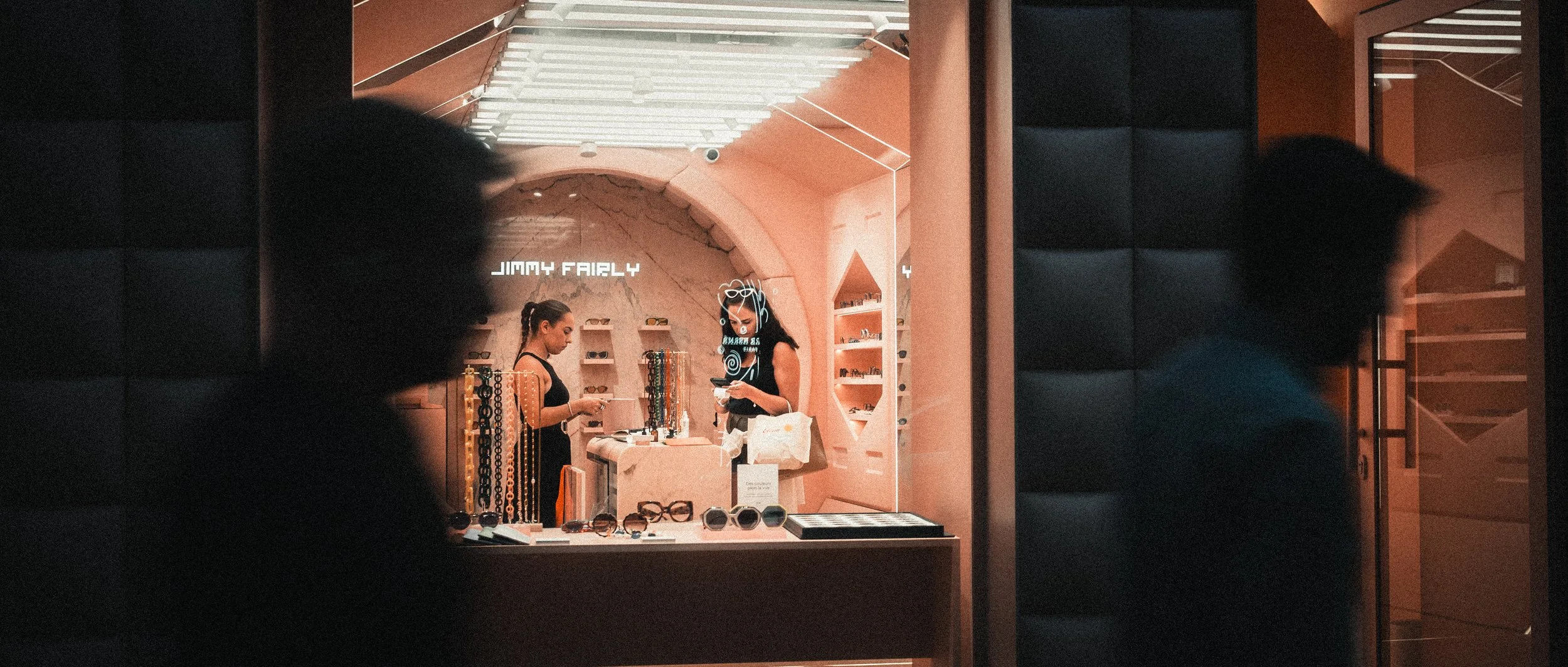 Deux femmes achetant des lunettes dans une boutique. La boutique porte un nom « Jimmy Fairly » et a une décoration en rose avec des étagères de lunettes en arrière-plan.