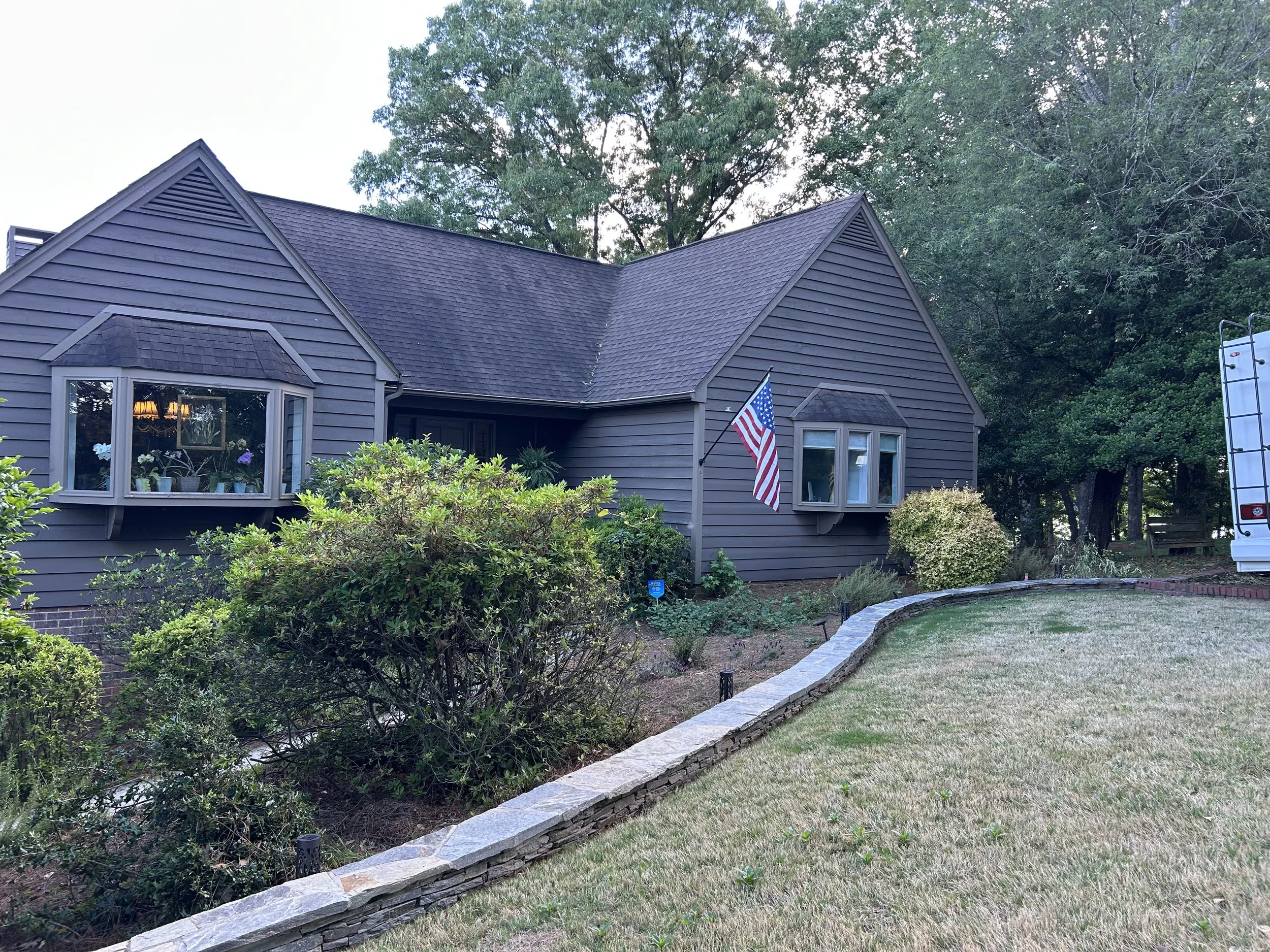 Gray house with two bay windows, one with flowers inside, American flag in front yard, landscaped lawn with stone border, trees in background.
