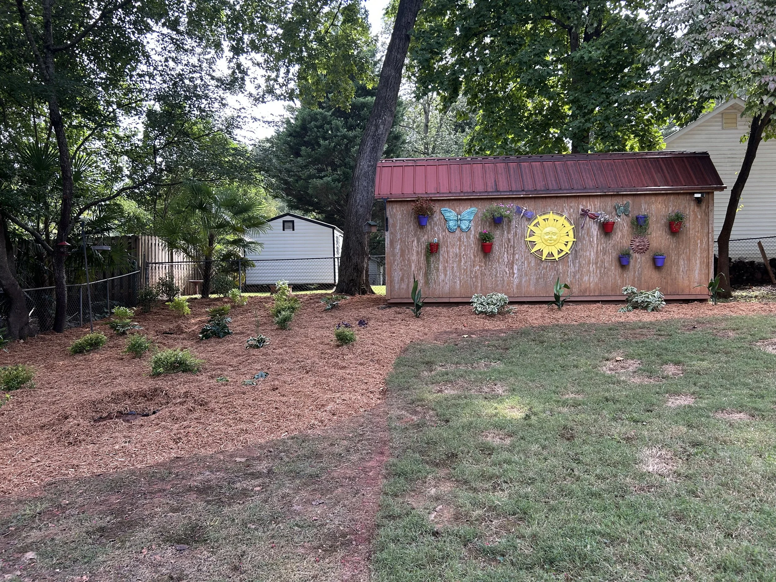 Backyard garden with small plants and flowers, a shed with decorative garden ornaments including a sun face, butterflies, and flower pots, surrounded by trees and fencing.