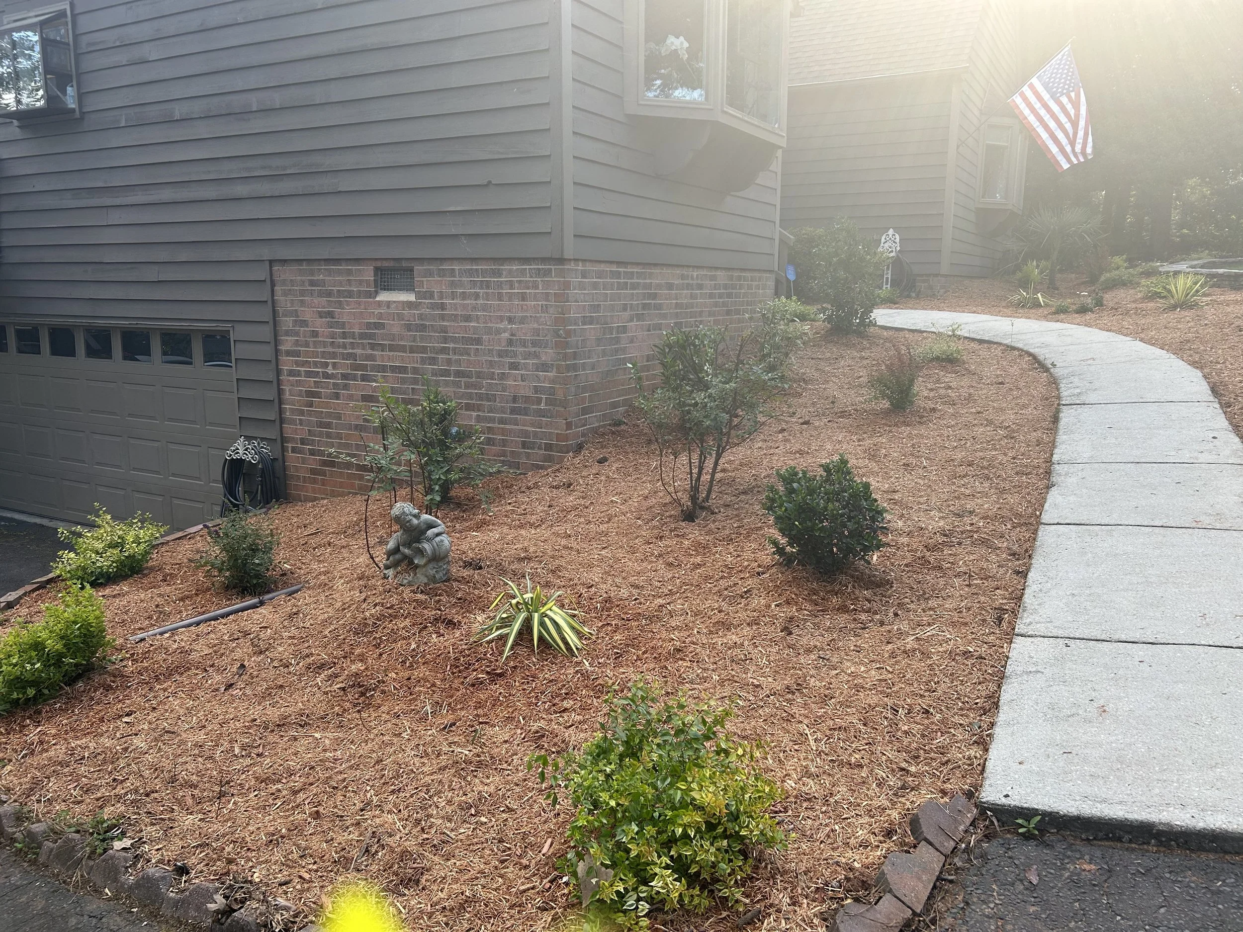 Front yard with a curved concrete walkway, small bushes, plants, and a garden ornament of a sitting frog, in front of a house with grey siding, brick foundation, American flag, and bay window.