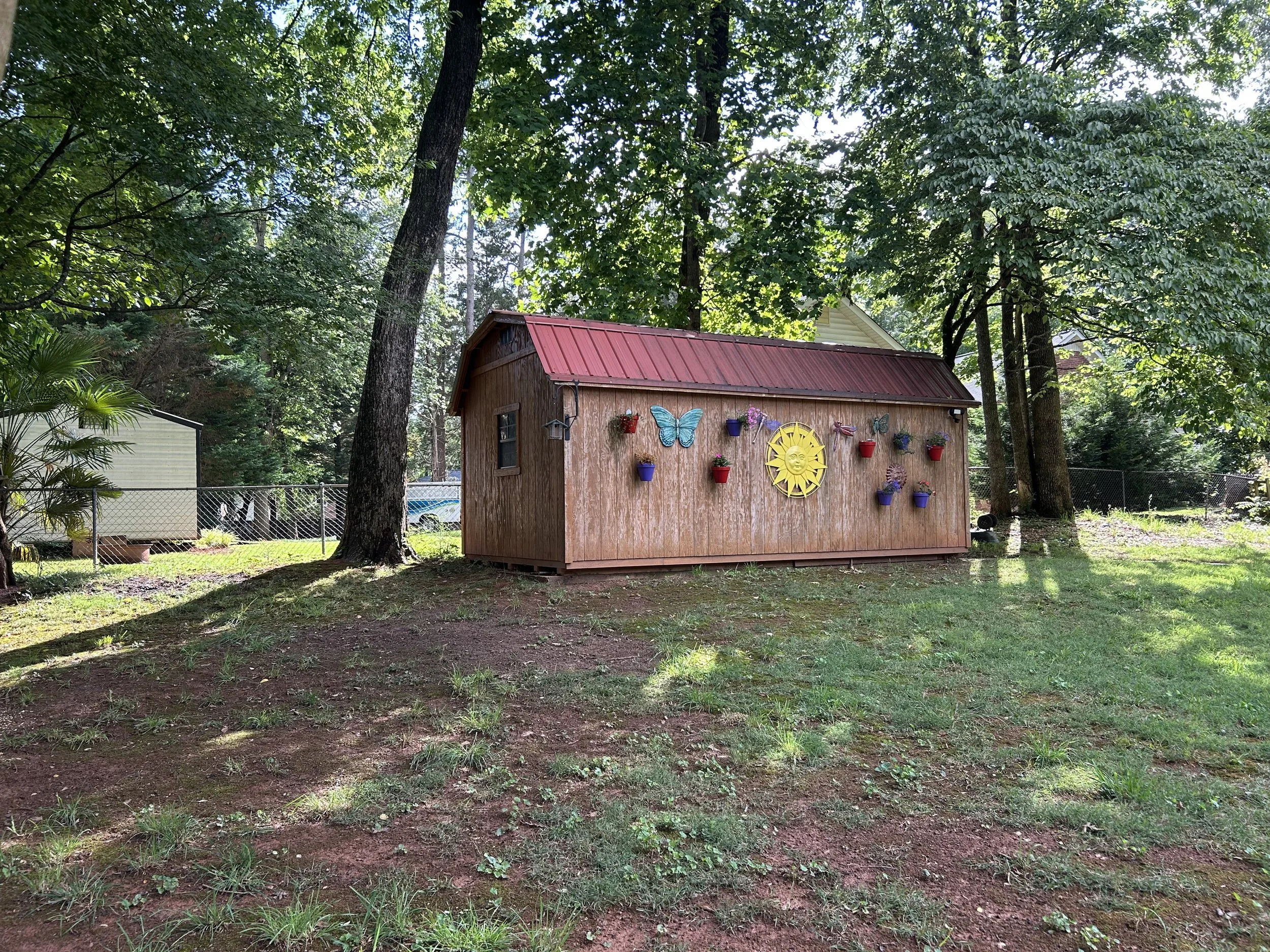 A small wooden shed decorated with colorful flower pots, a sun face, butterfly, and bird ornaments, surrounded by trees in a backyard.