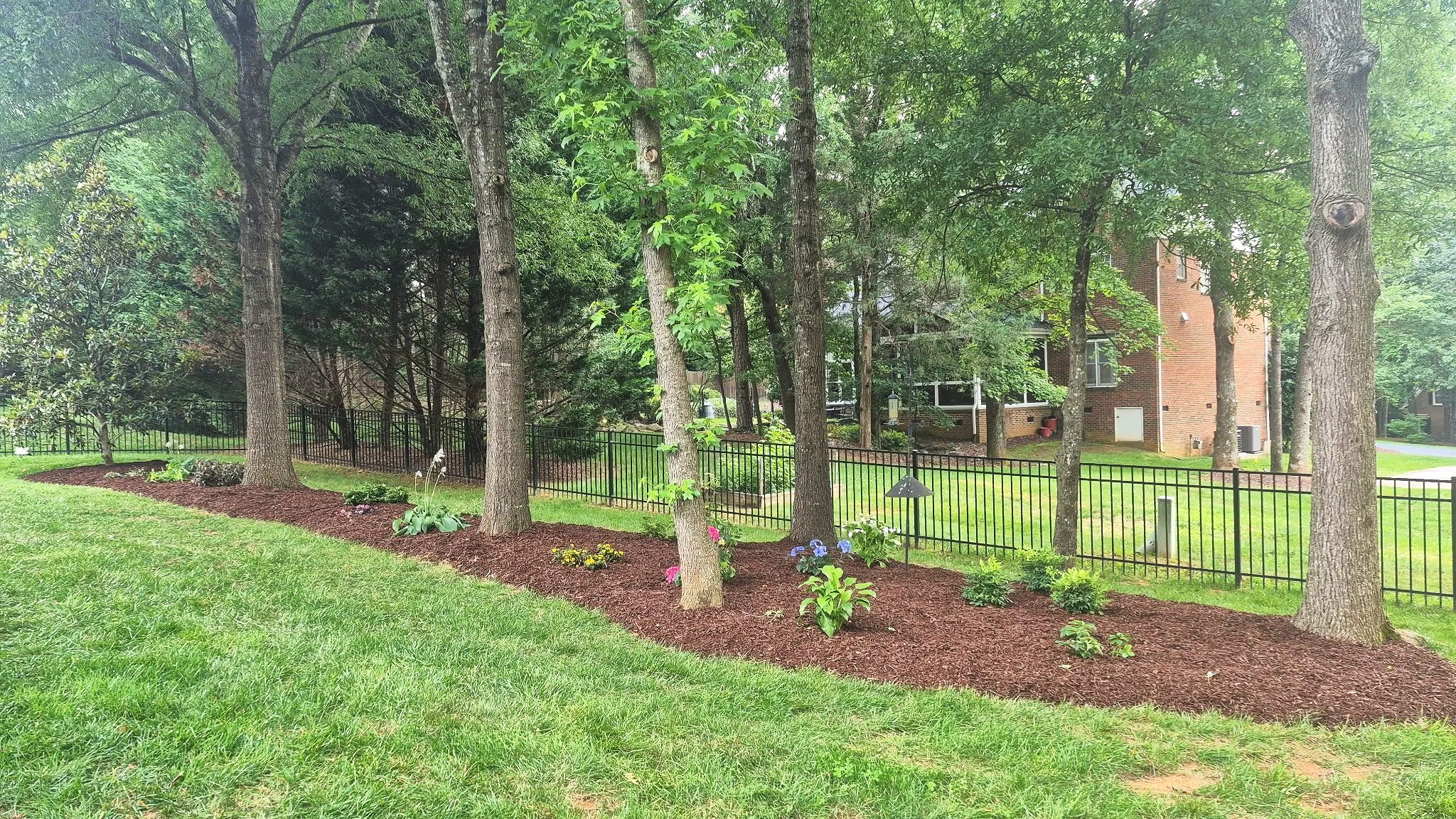 A landscaped yard with a row of trees, colorful flowers, and a black metal fence, with a multi-story brick building in the background.