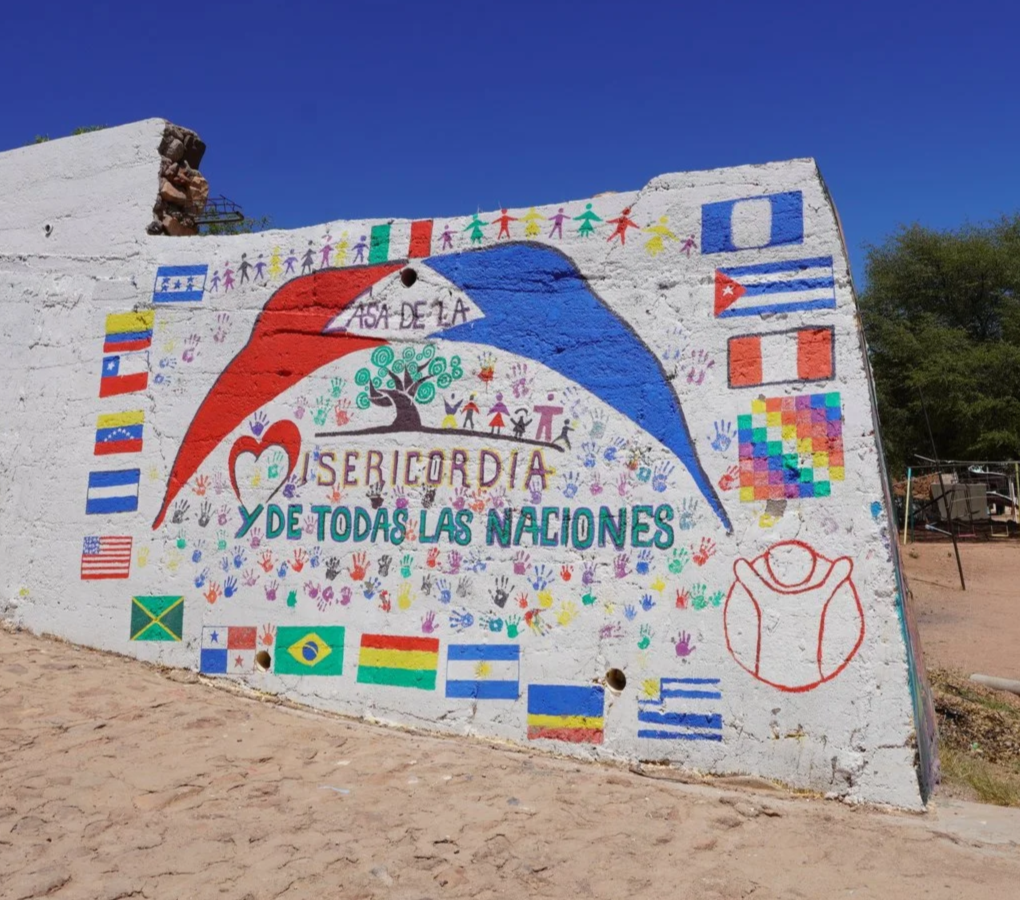 Colorful mural on a white wall with flags of various countries, a heart, a tree, and children holding hands. It features a red, blue, and white design in the shape of a hat and includes the words "Seriordia y de todas las naciones" and "Asia De La".