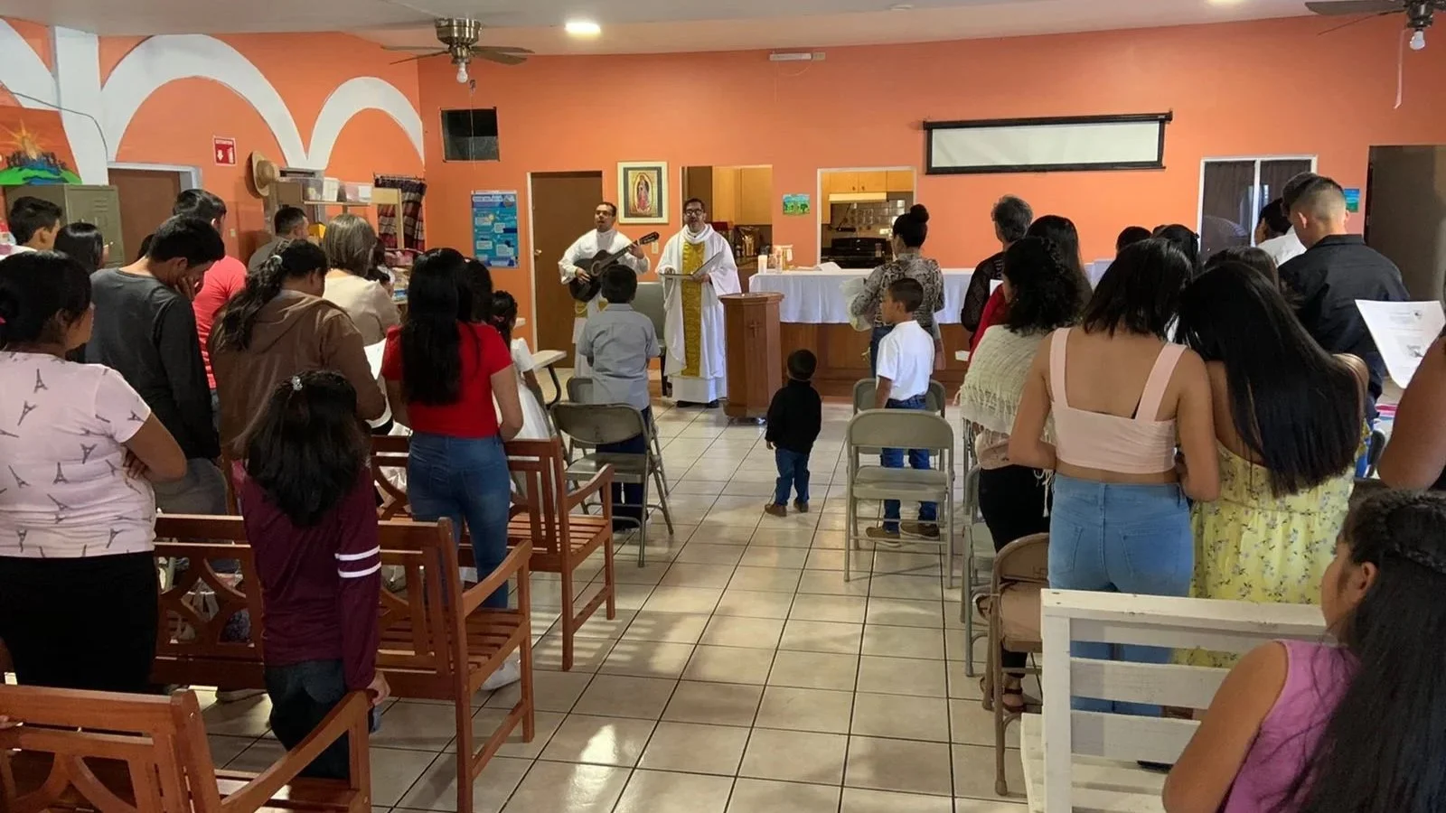 A church service with a congregation participating in prayer, with two priests in white and gold robes, one playing a guitar, standing at the altar in a room with orange walls and tiled floor.