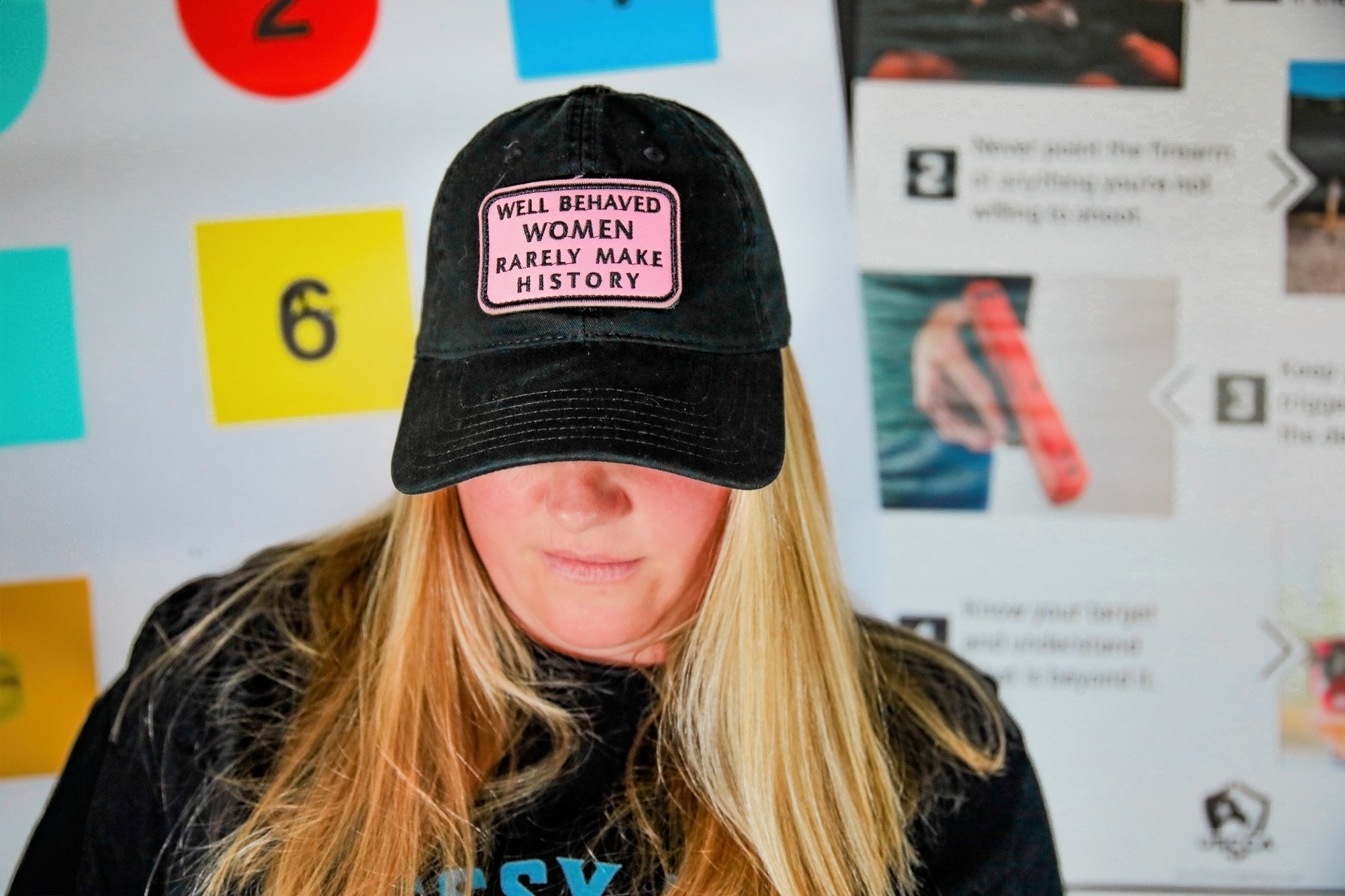A woman wearing a black cap with a pink patch that reads, "WELL BEHAVED WOMEN RARELY MAKE HISTORY." She has long blonde hair and is wearing a black top with blue text. The background features various colorful posters and signs on a wall.