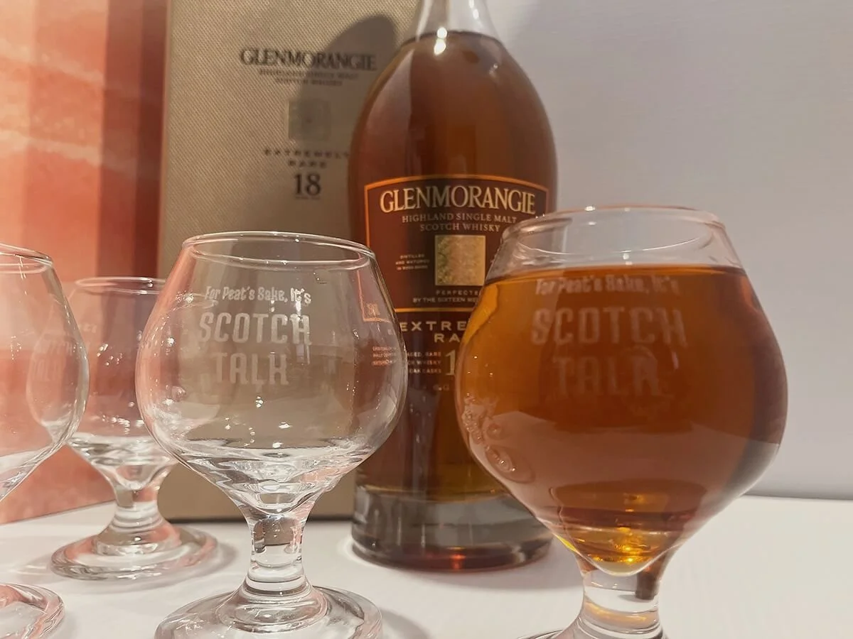 Two scotch glasses with custom branded etching on the front. One of them is full of scotch. There is a scotch whiskey bottle in the background.
