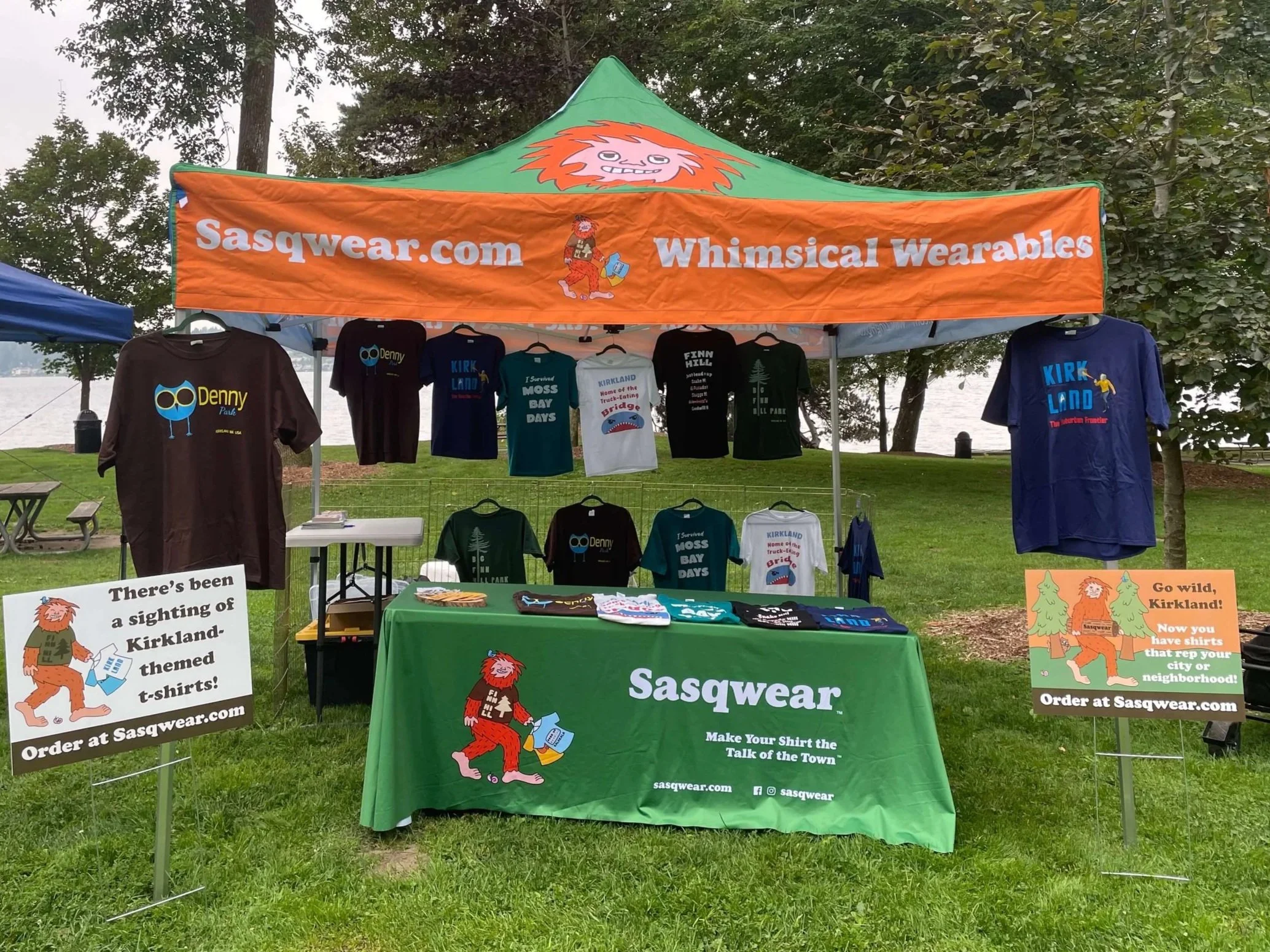 A canopy set up at an event outside. The canopy has several custom branded t-shirts hanging inside of it, along with a branded tablecloth and yard signs.