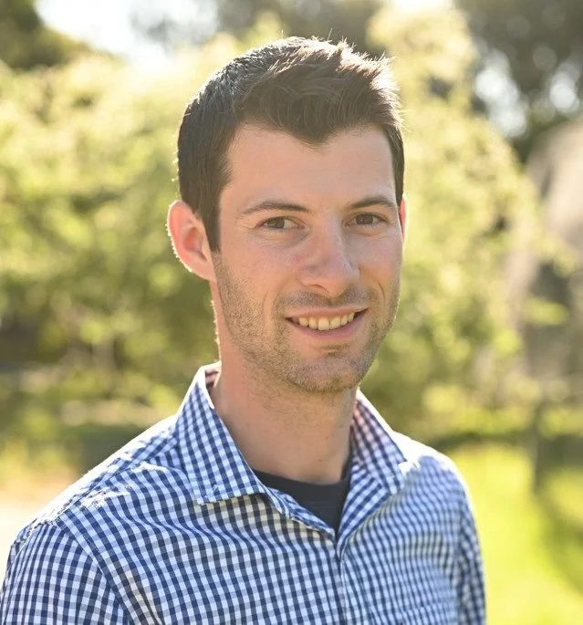A young man with short dark hair, wearing a blue checkered shirt, smiling outdoors with a blurred background of trees and sunlight.