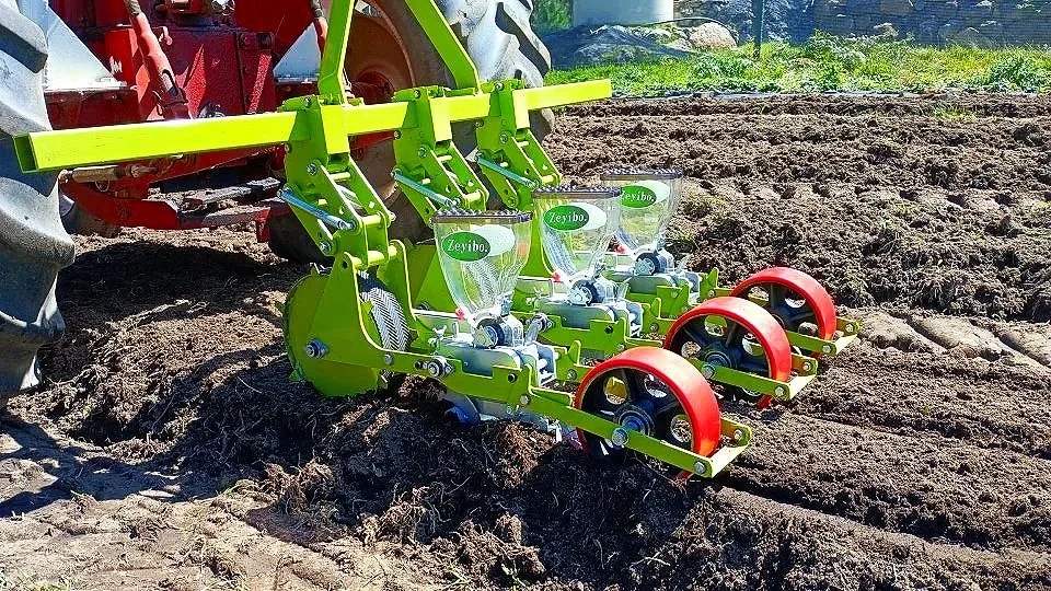 A close-up of a specialized piece of farming equipment attached to a tractor, designed for planting seeds in soil. The equipment has green and red components and clear seed containers labeled "Zeyibo." The machinery operates on a row of freshly tilled soil.