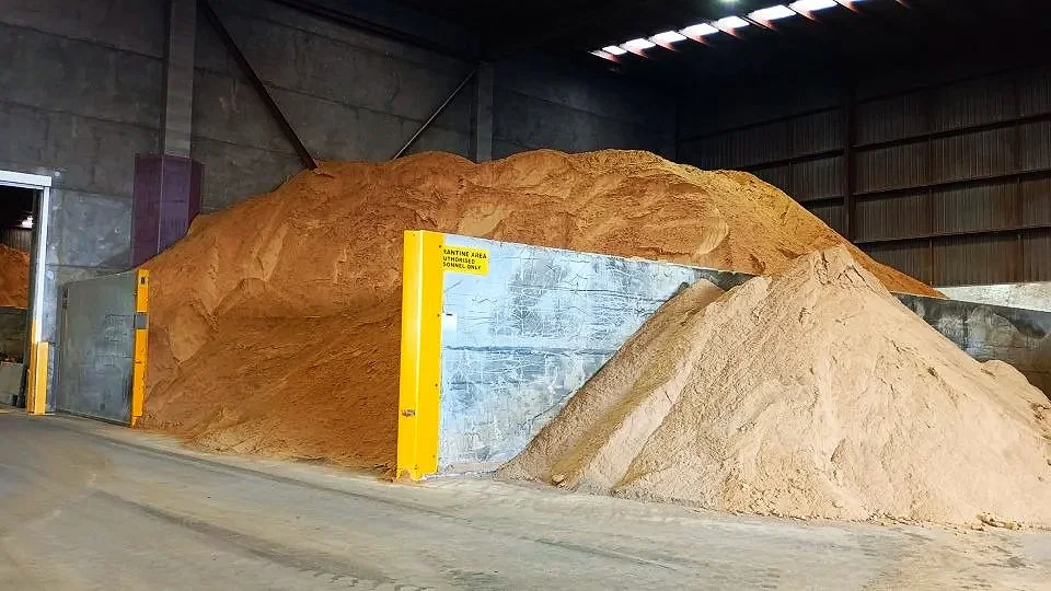 Large pile of sand inside an industrial warehouse with a smaller pile in front and yellow safety barriers along the side.