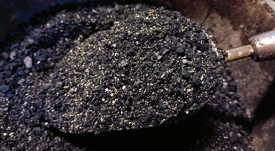 Close-up of a metal spoon filled with black and silver granular material, surrounded by similar granular material.