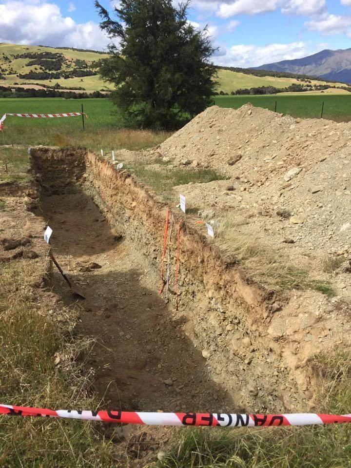 A construction site with a deep, narrow trench in the ground, surrounded by dirt mounds, with caution tape marking the area and a rural landscape with green fields, trees, and mountains in the background.