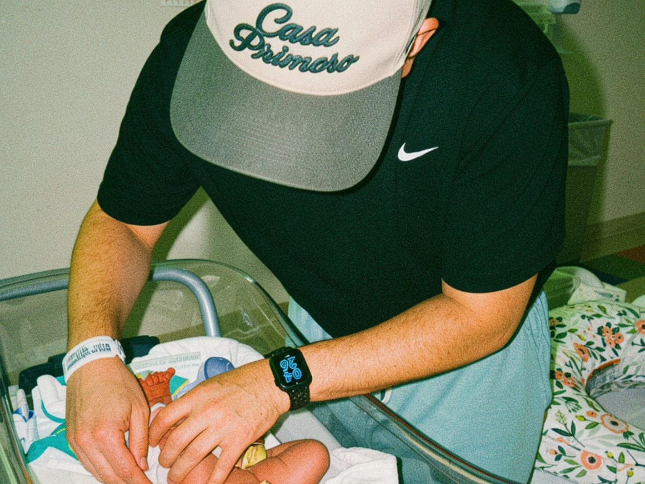 Person wearing a cap with the words "Casa Primero", a black Nike t-shirt, and a smartwatch, attending to a newborn in a hospital bassinet.