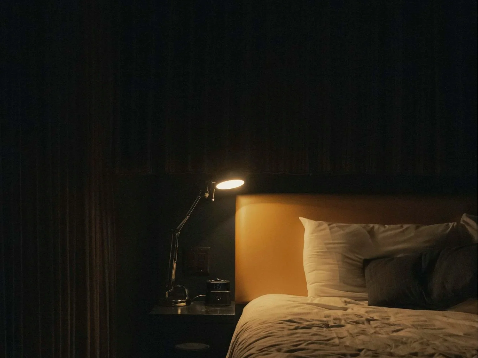 A dimly lit bedroom with a bed, a nightstand, a digital alarm clock, and a bedside lamp. The bed has white pillows and a beige headboard. The lamp provides soft lighting against dark curtains and wall.
