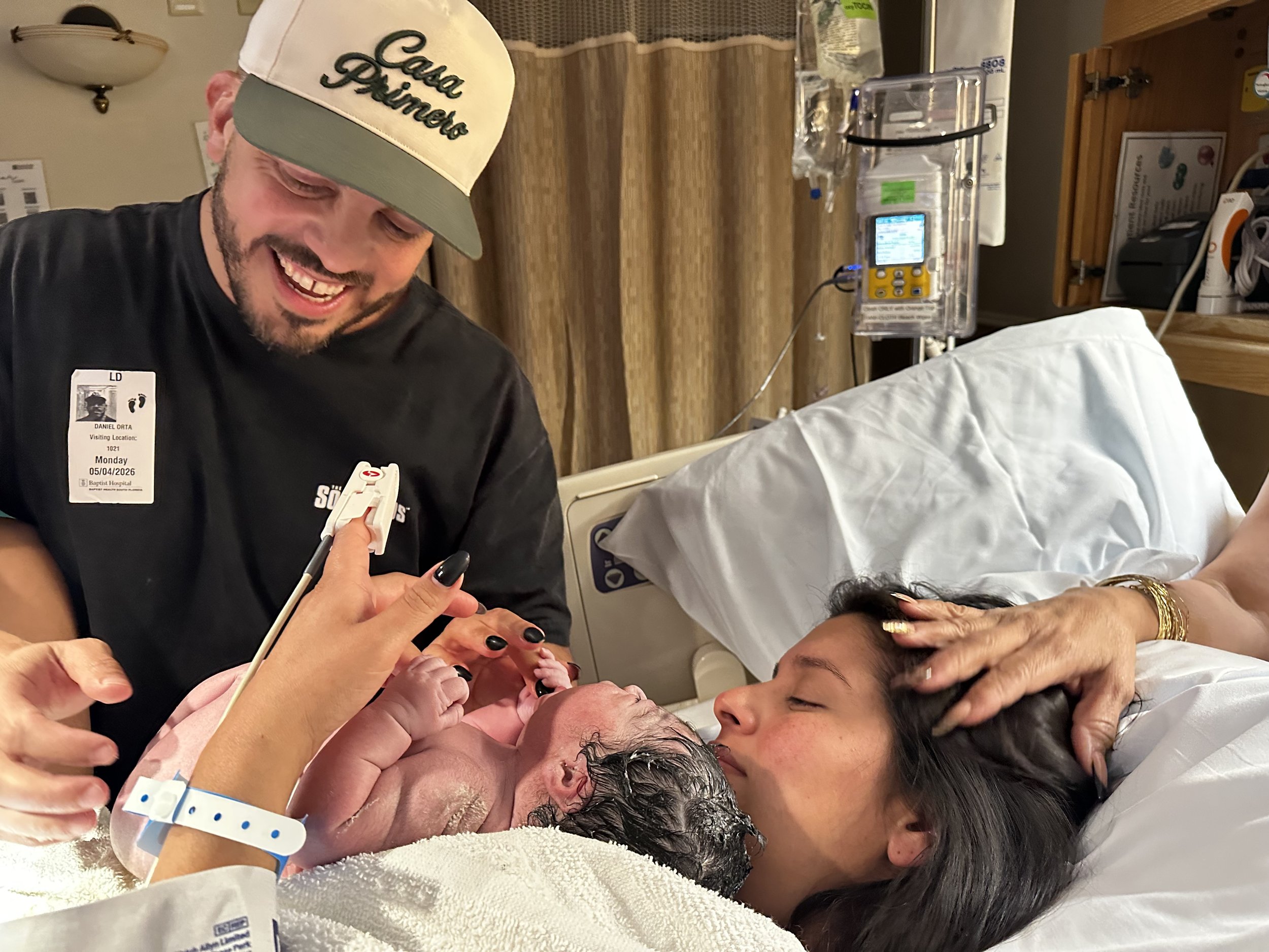 A man wearing a cap with 'Casa Primero' embroidered on it, smiling, is holding a newborn baby. The baby is being cleaned or examined by healthcare staff with gloves. The scene appears to be in a hospital room with medical equipment and a patient bed.