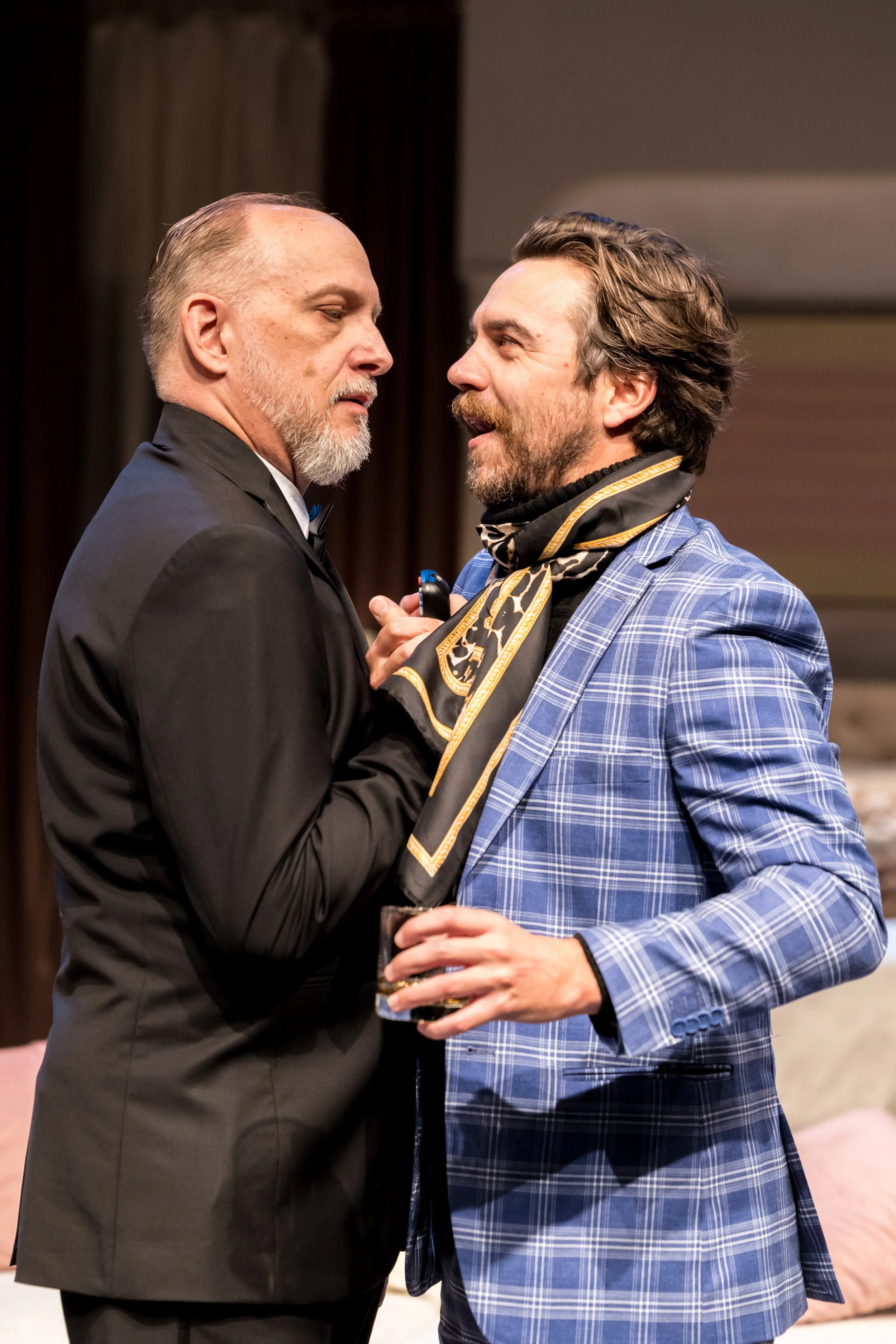 Two men standing close together, one in a black tuxedo with a grey beard and the other in a blue plaid blazer with a scarf, having a conversation during a social event, with one holding a glass of drink.