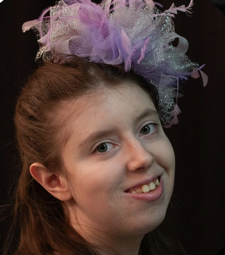 Young girl with reddish-brown hair wearing a colorful, fluffy, and glittery headpiece, smiling with a black background.