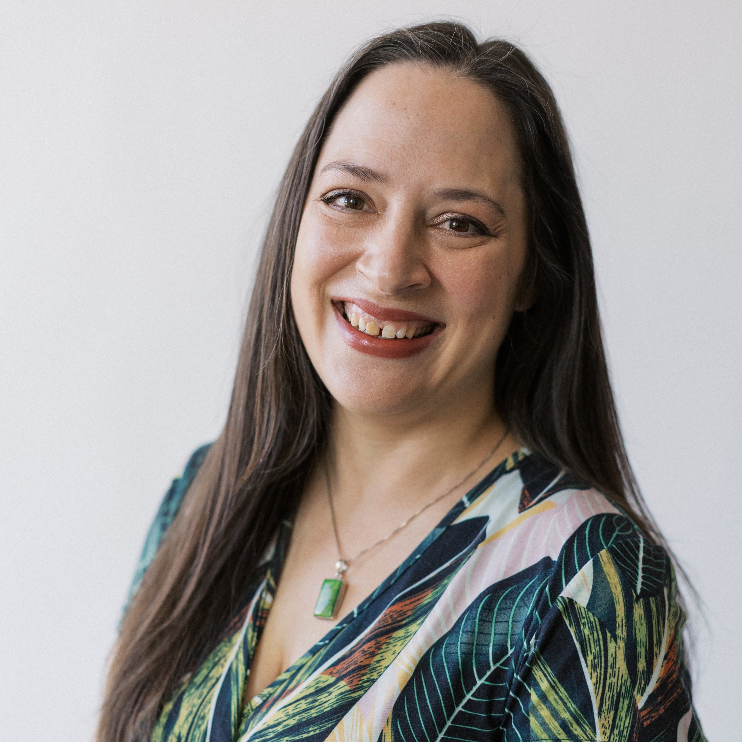 A woman with long dark hair smiling, wearing a colorful patterned blouse and a green pendant necklace, standing against a plain white background.