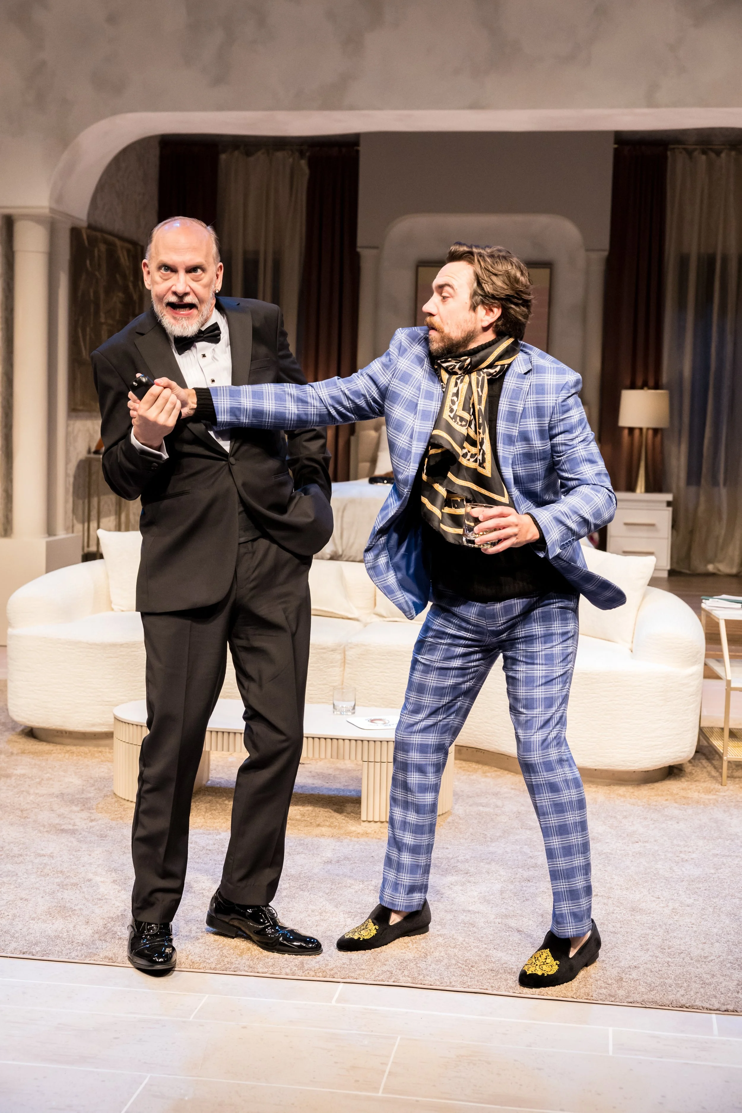 A man in a black tuxedo and bowtie is holding a remote and looks surprised while another man in a blue plaid suit and scarf appears to be pulling him or gesturing toward him during a theatrical performance in a living room setting.
