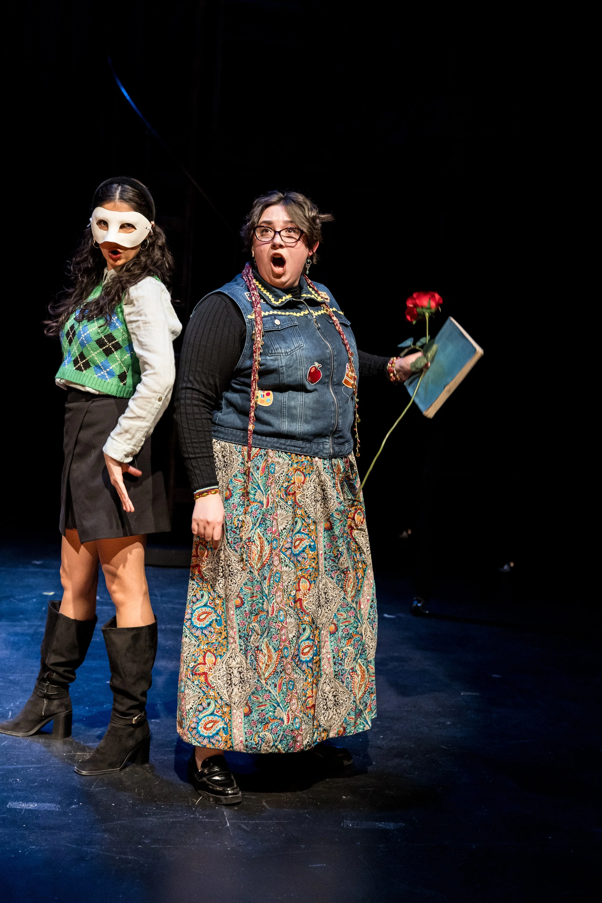 Two women standing on stage, one wearing a white mask and a green argyle sweater vest, and the other holding a red rose, a book, and appearing to be speaking passionately.