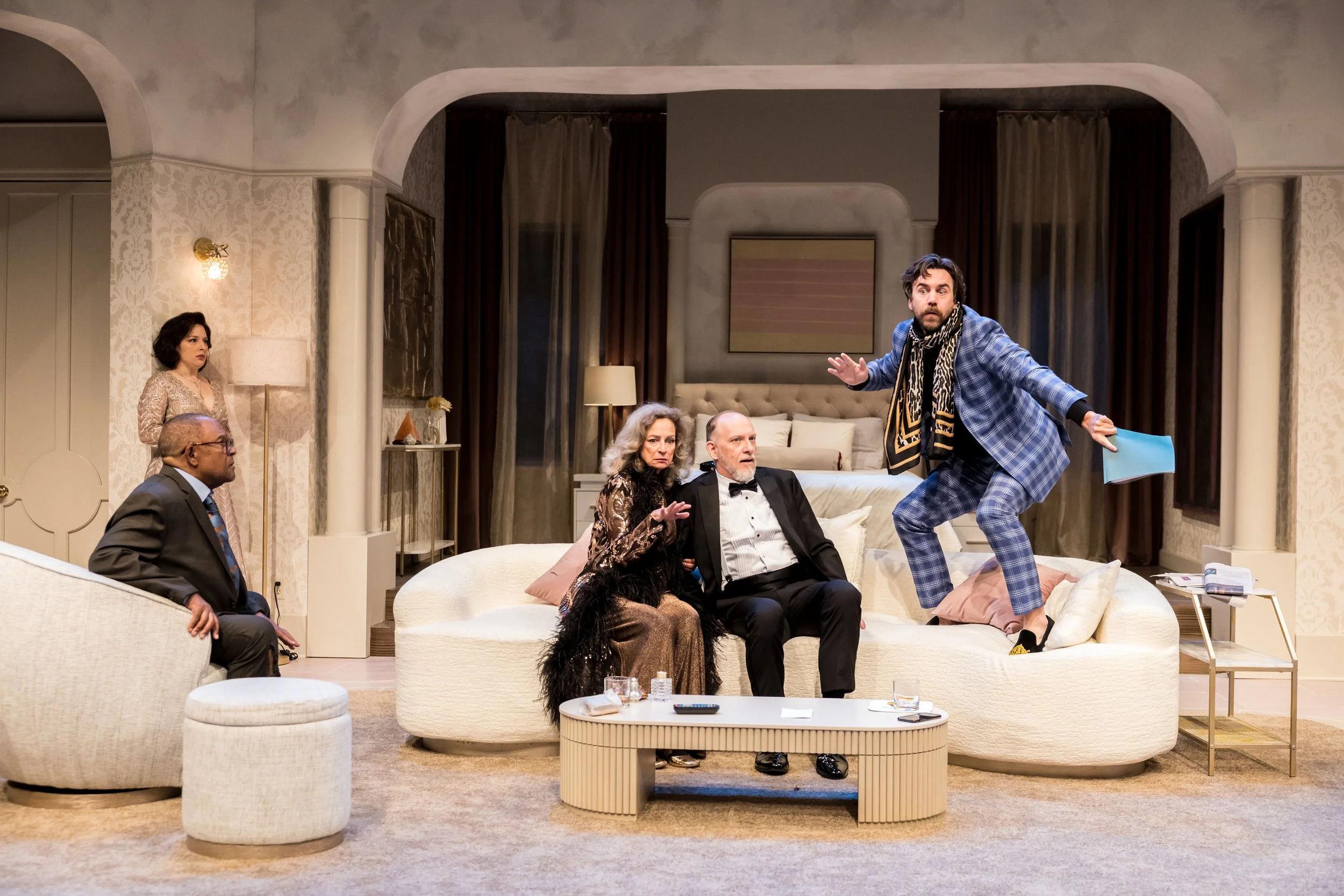 People acting in a play or skit in a living room setting. One person is standing on a sofa, gesturing with hands, while the others are seated and watching. The scene is staged with furniture and decor resembling a modern apartment.