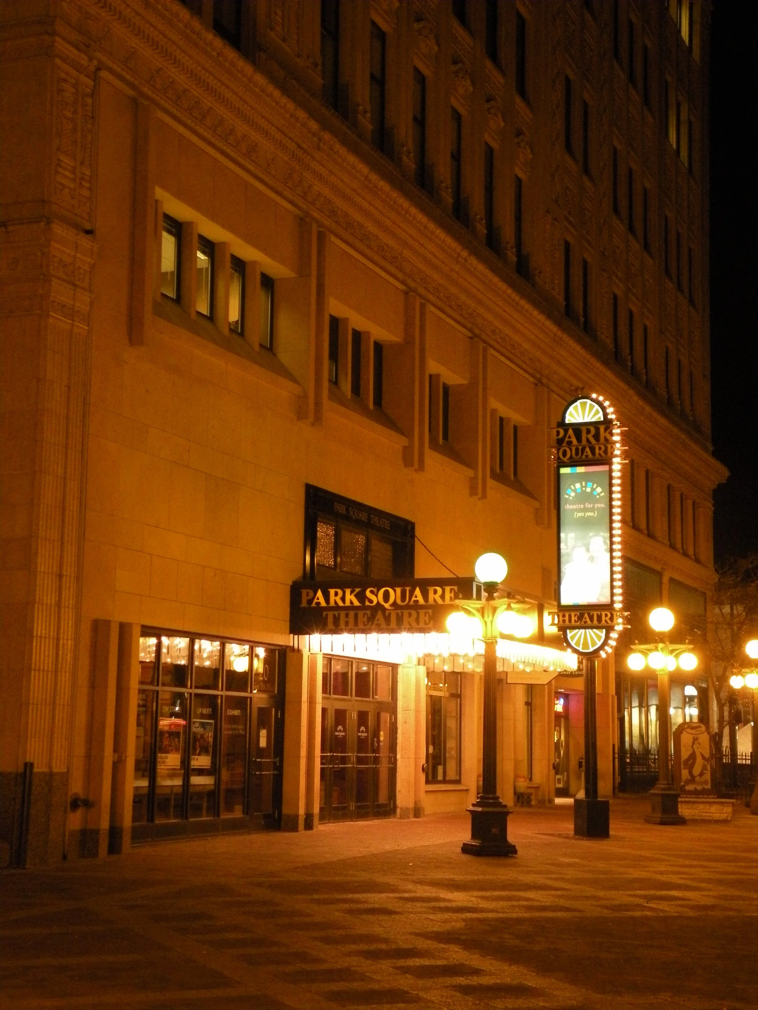 Night view of the exterior of Park Square Theatre with illuminated sign and street lamps.