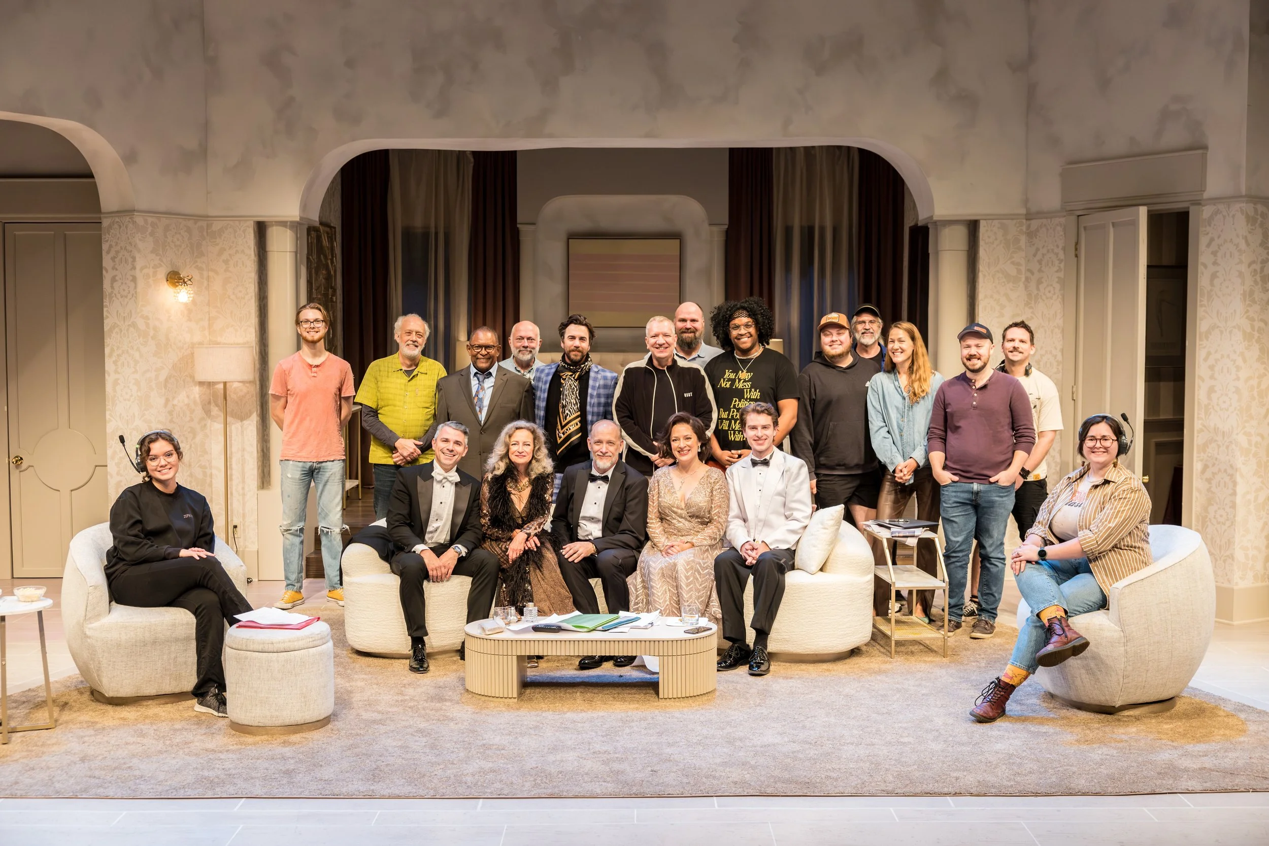 A group of people posing together on a stage with elegant decor, including a beige couch, chairs, and a coffee table, in a well-lit room.