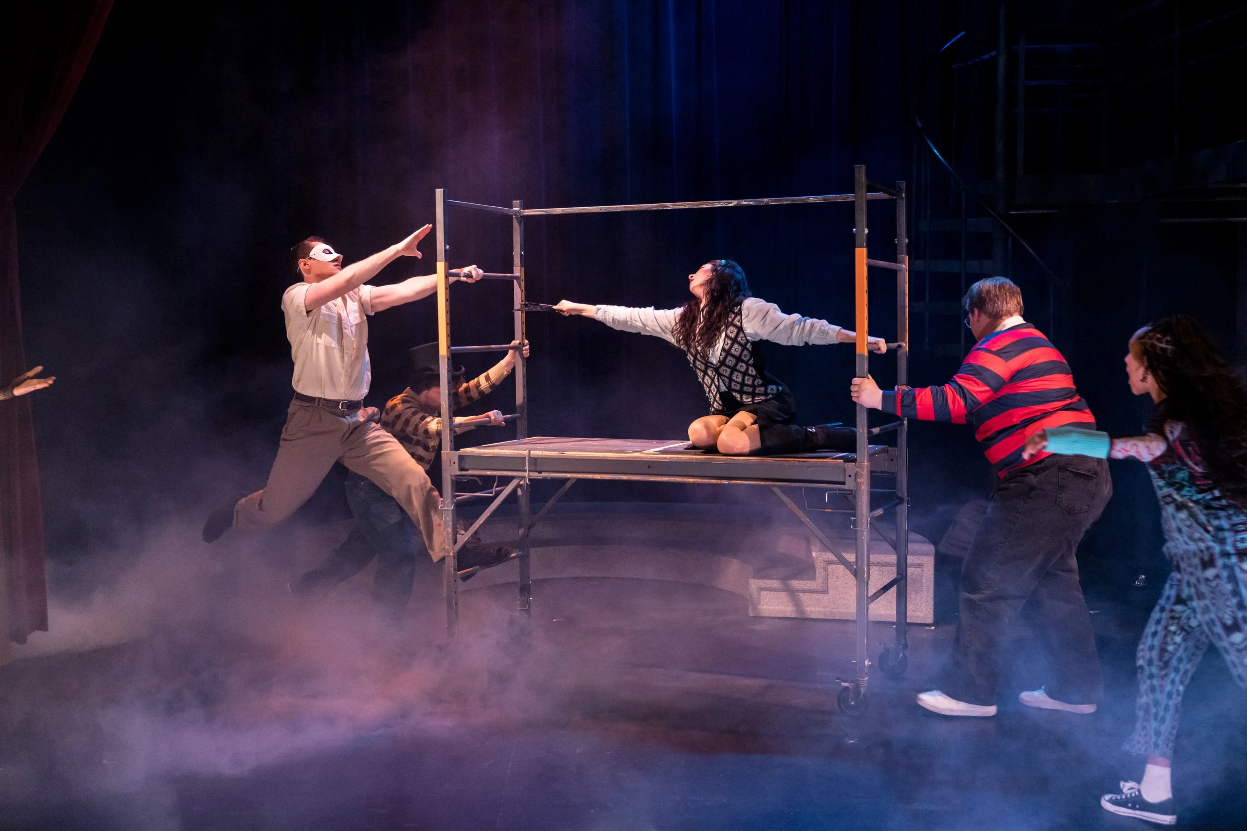 The image shows a theatrical performance with six actors on stage, one woman sitting cross-legged on a lift platform in the center, extending her hand, and five others surrounding her, some pulling or pushing the platform. The stage has a smoky atmos