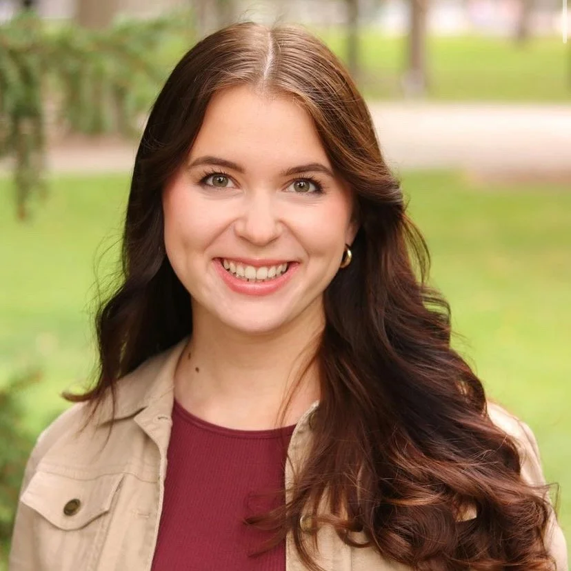 A young woman with long brown hair smiling outdoors with a grassy background.