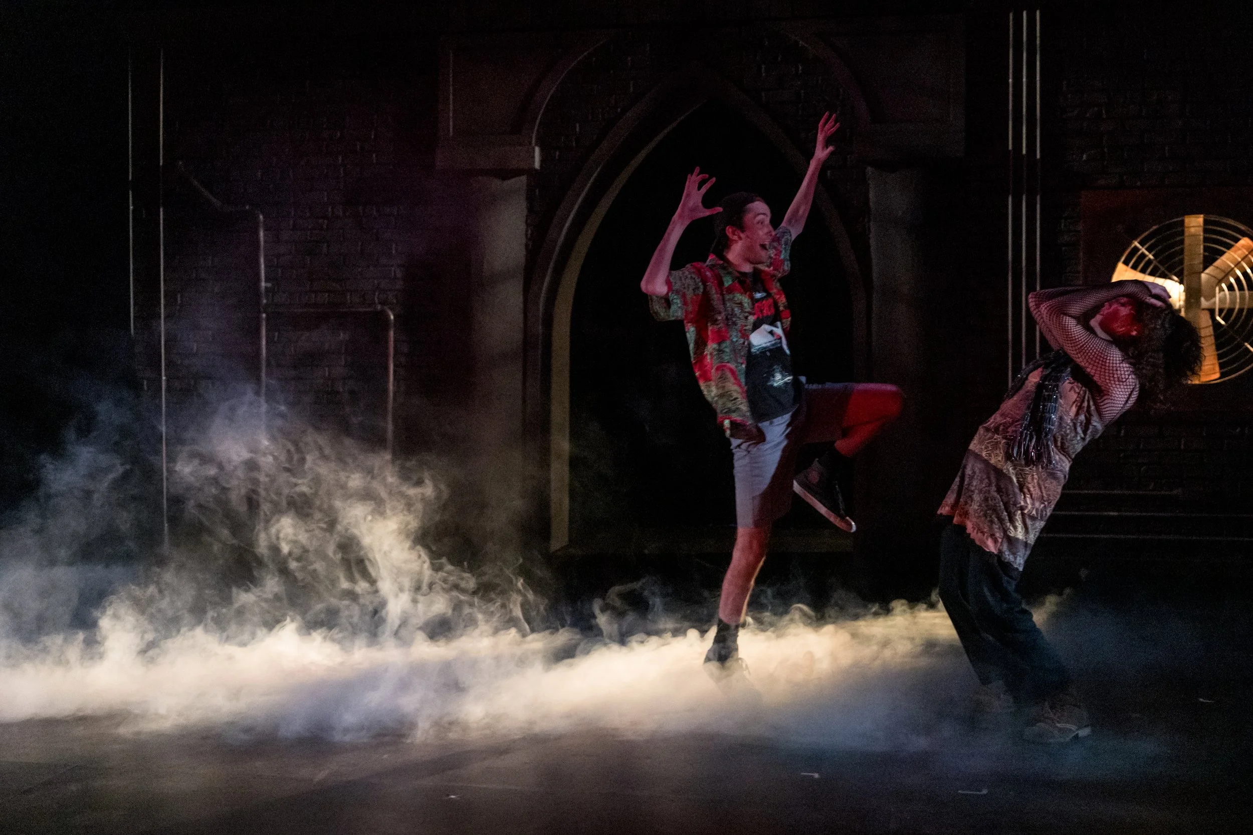 Two performers on a stage with smoke, one with arms raised and one leaning back, in a theatrical scene.