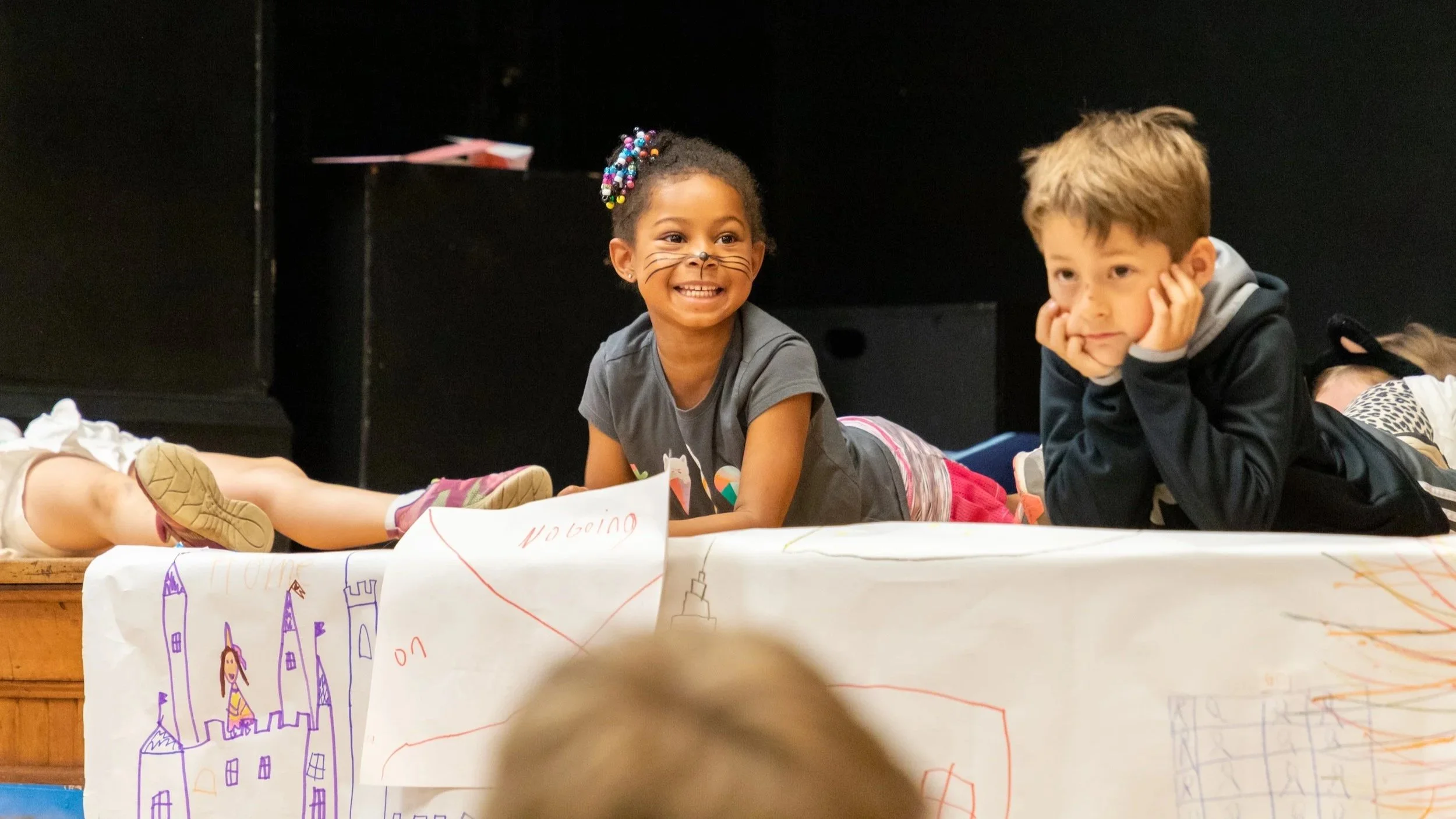 Two children lying on their stomachs on a stage, smiling and watching the audience, with drawings on a large paper covering the front of the stage.