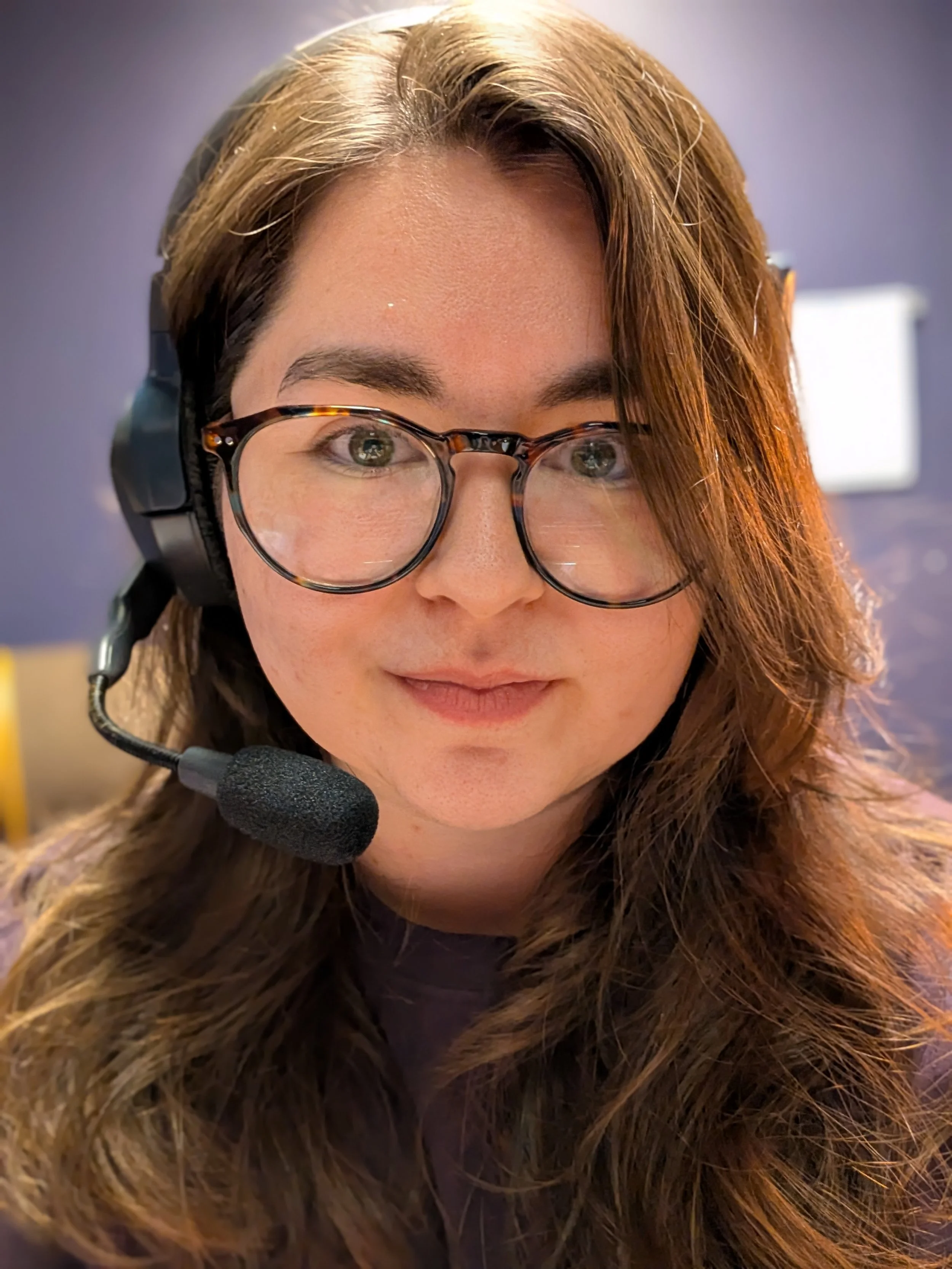 Close-up of a young woman with brown hair, glasses, and a slight smile, in a room with green walls and a mirror in the background.
