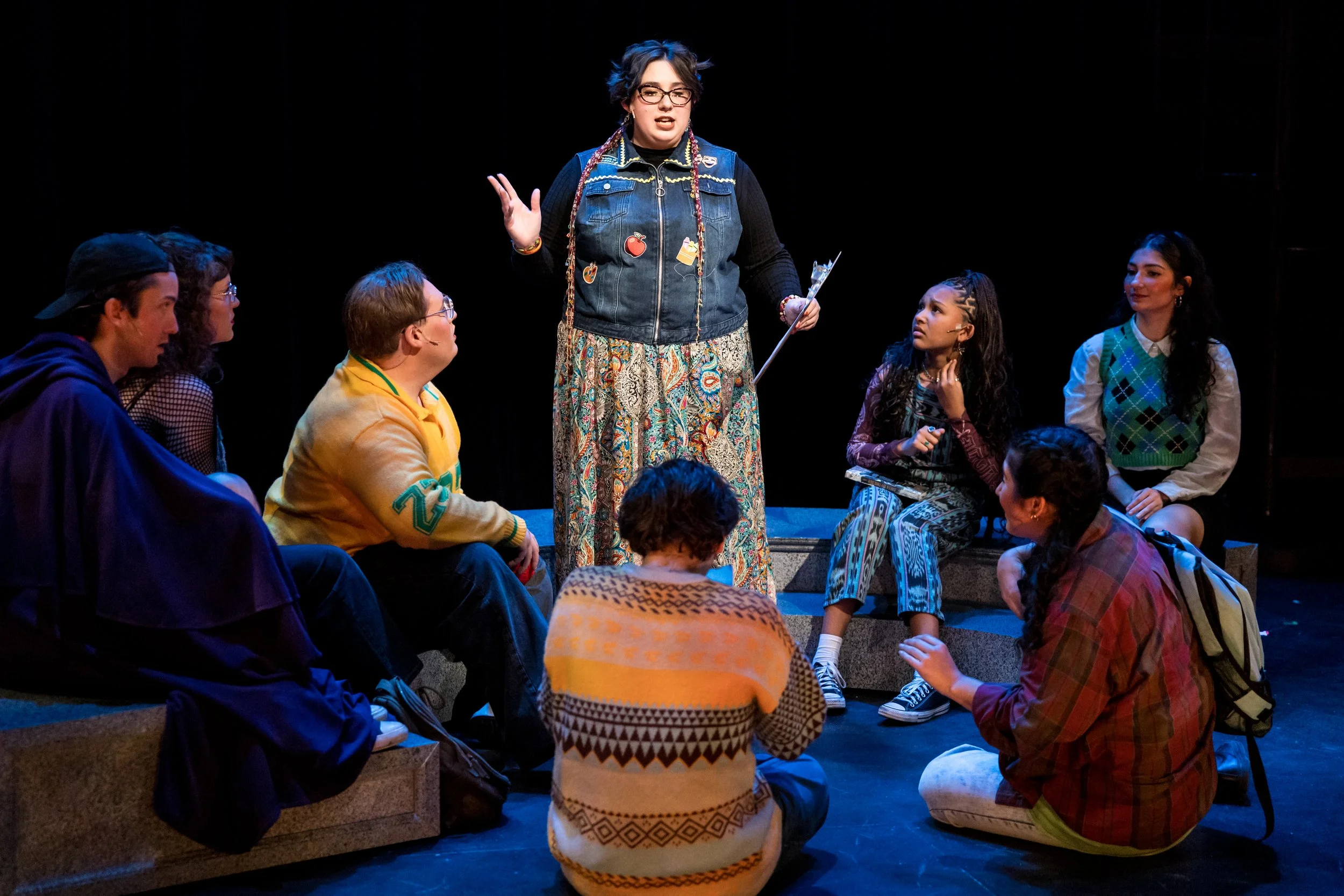 A young woman with glasses, colorful hair, a denim vest with patches, and patterned pants is speaking to a diverse group of young people seated around her on a stage. The group is listening attentively in a theatrical performance setting.