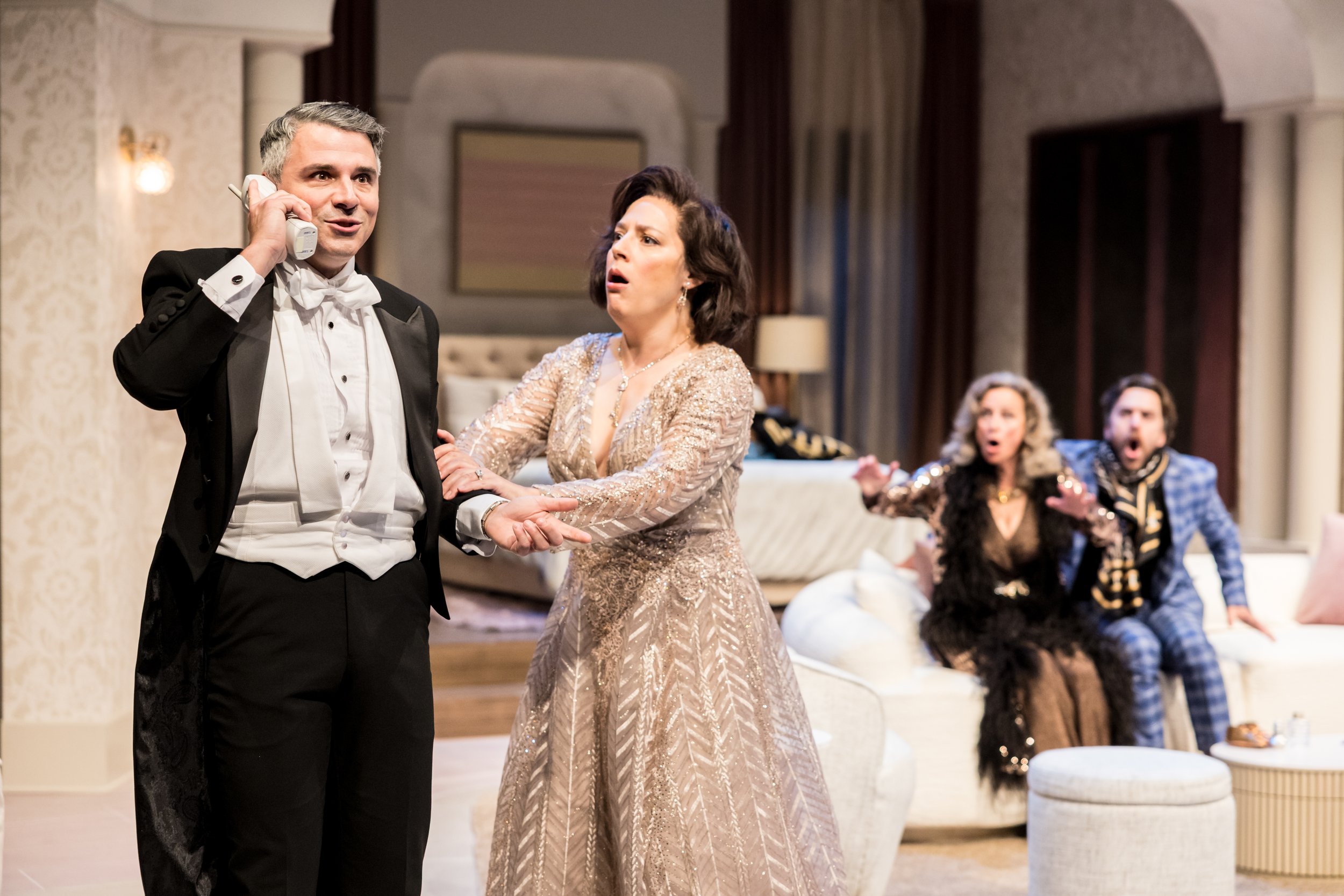 Four people on stage in a theatrical play. One man in tuxedo talking on the phone, a woman in a gold dress looking shocked, and two other people on the couch in the background reacting with surprise.