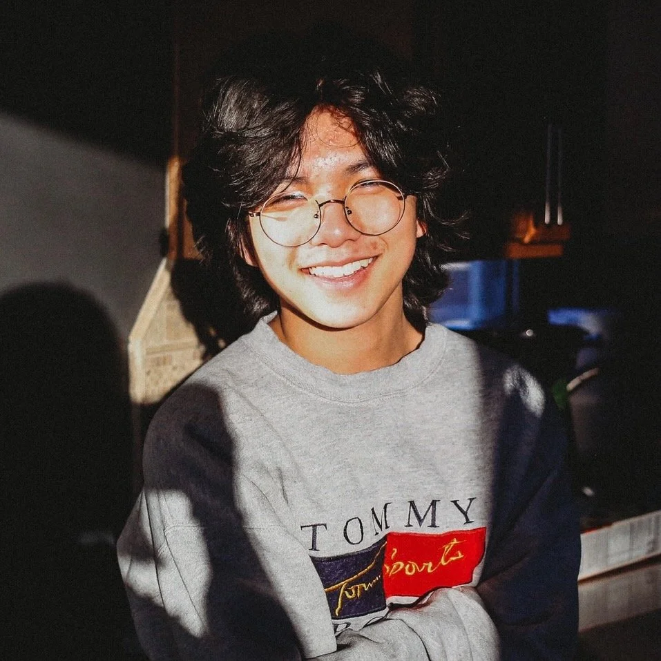 A young man with curly black hair and glasses smiling in a dimly lit room, wearing a gray Tommy sweatshirt.